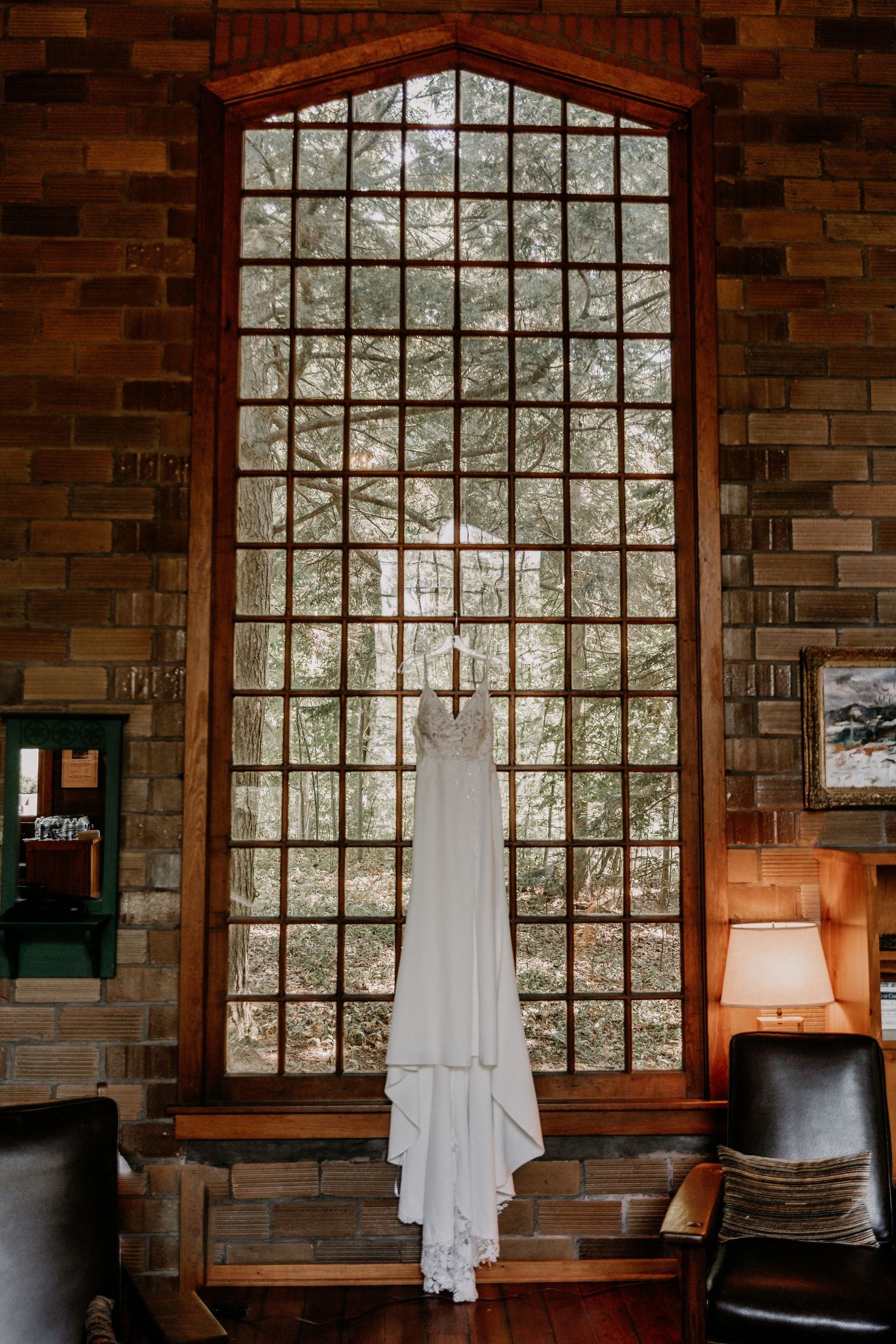A white wedding dress hangs elegantly against a large arched window with grid panes in a brick-walled room.