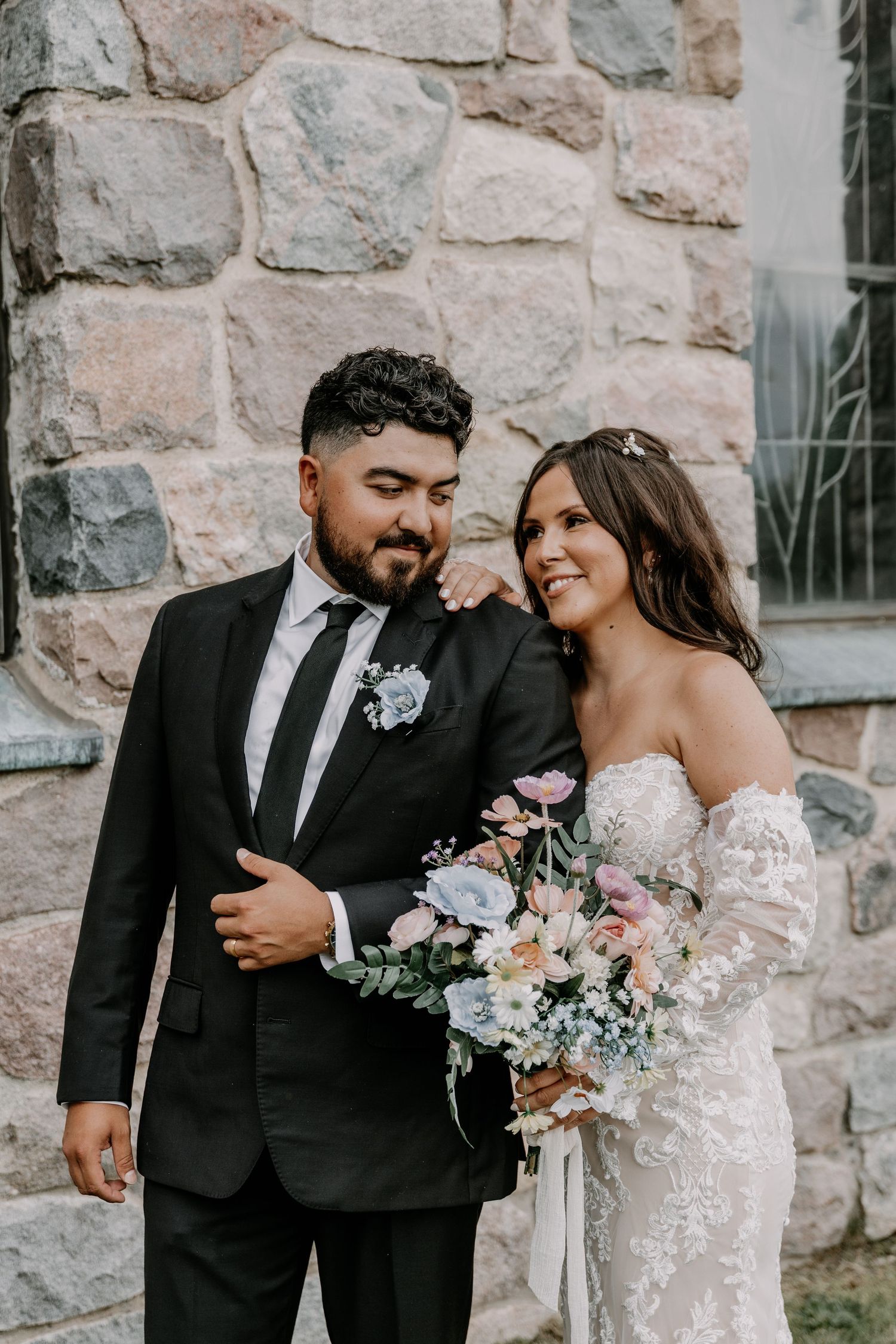 A romantic wedding couple dressed in formal attire standing by a rustic stone wall with a beautiful floral bouquet.