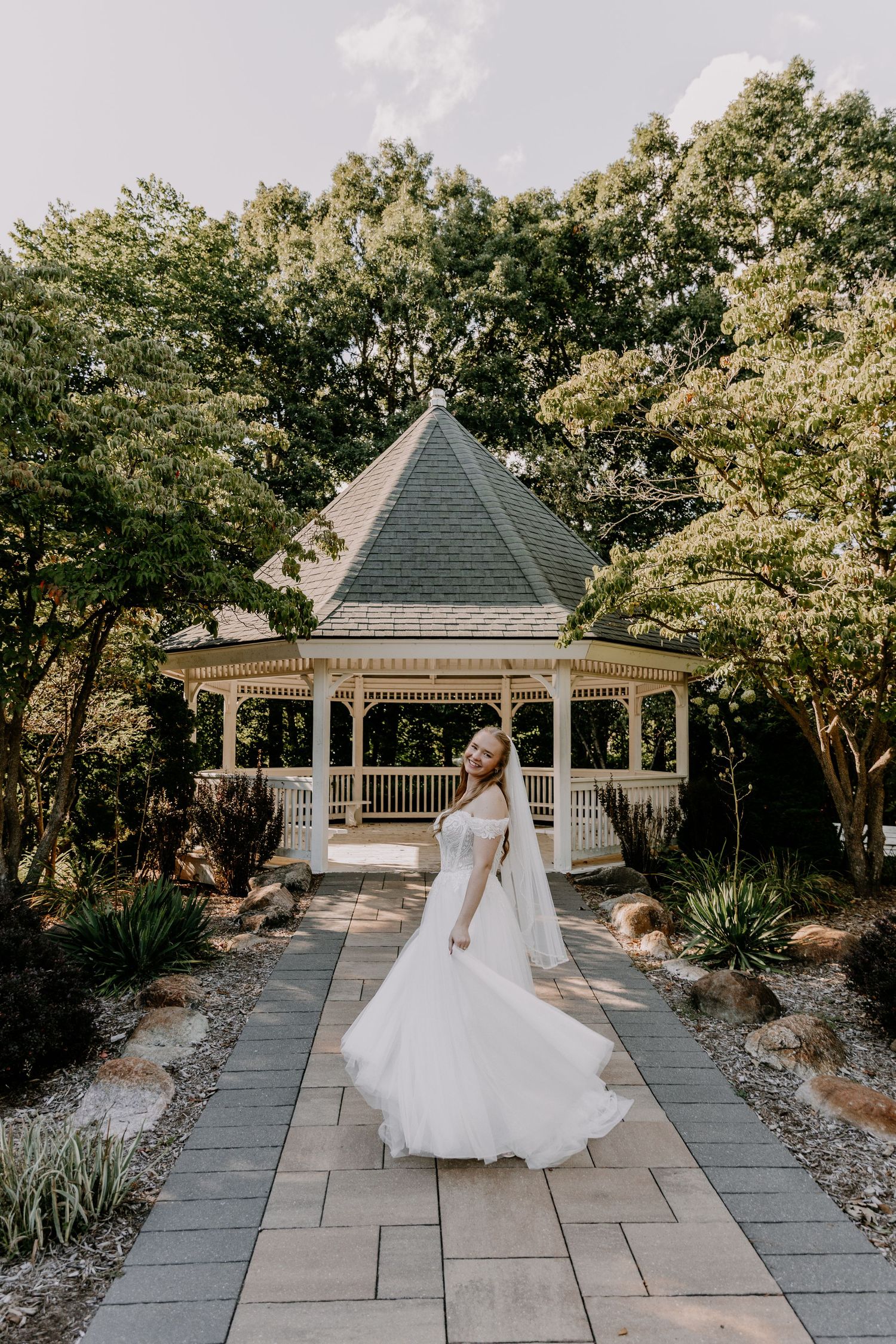 Bride in flowing white off-shoulder gown twirling on stone path before elegant white gazebo surrounded by lush greenery.