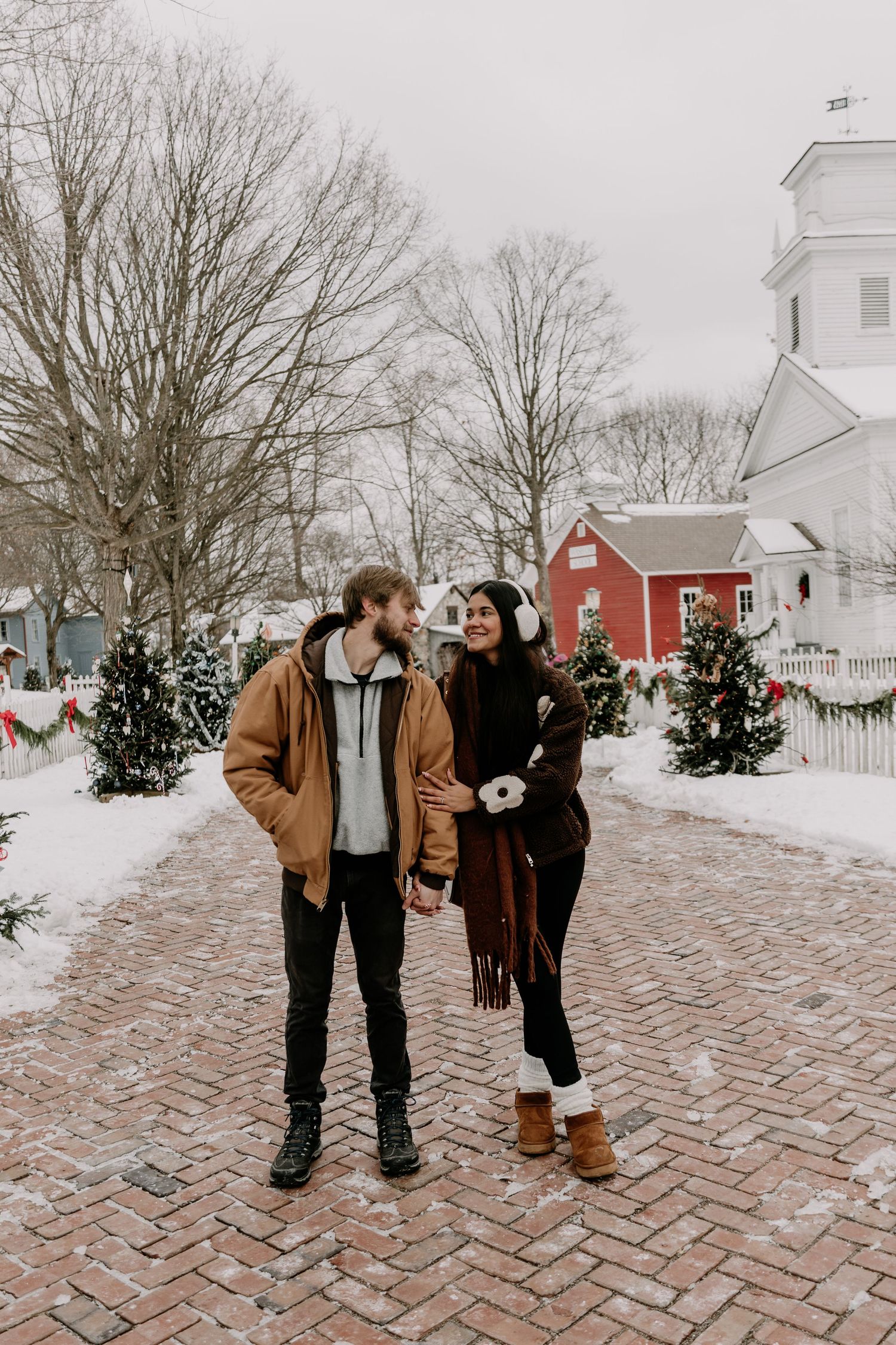 A couple shares intimate moments on a snow-covered brick pathway with red buildings in the background.