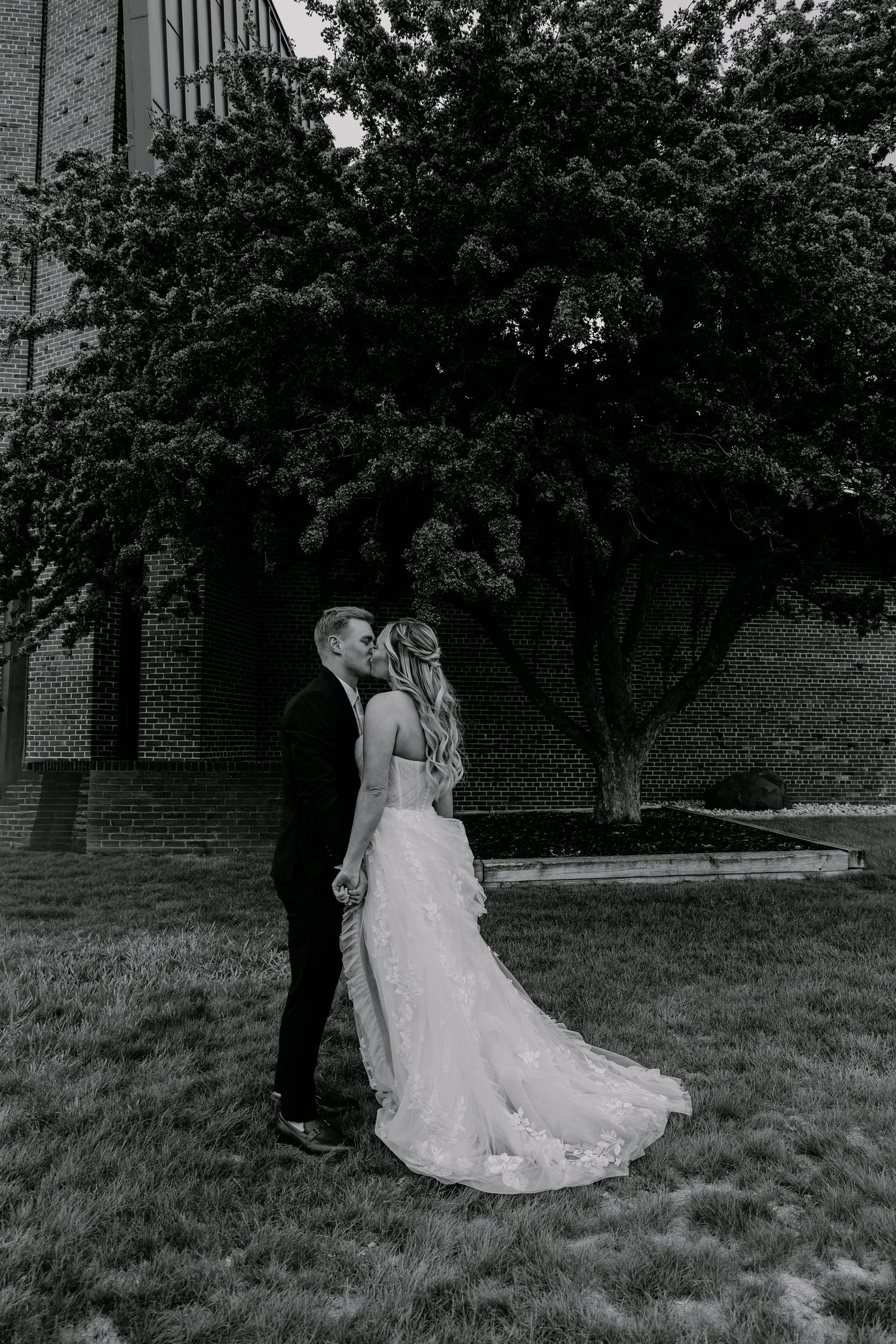 Black and white photo of a bride and groom sharing a kiss outdoors near a brick building and large tree.