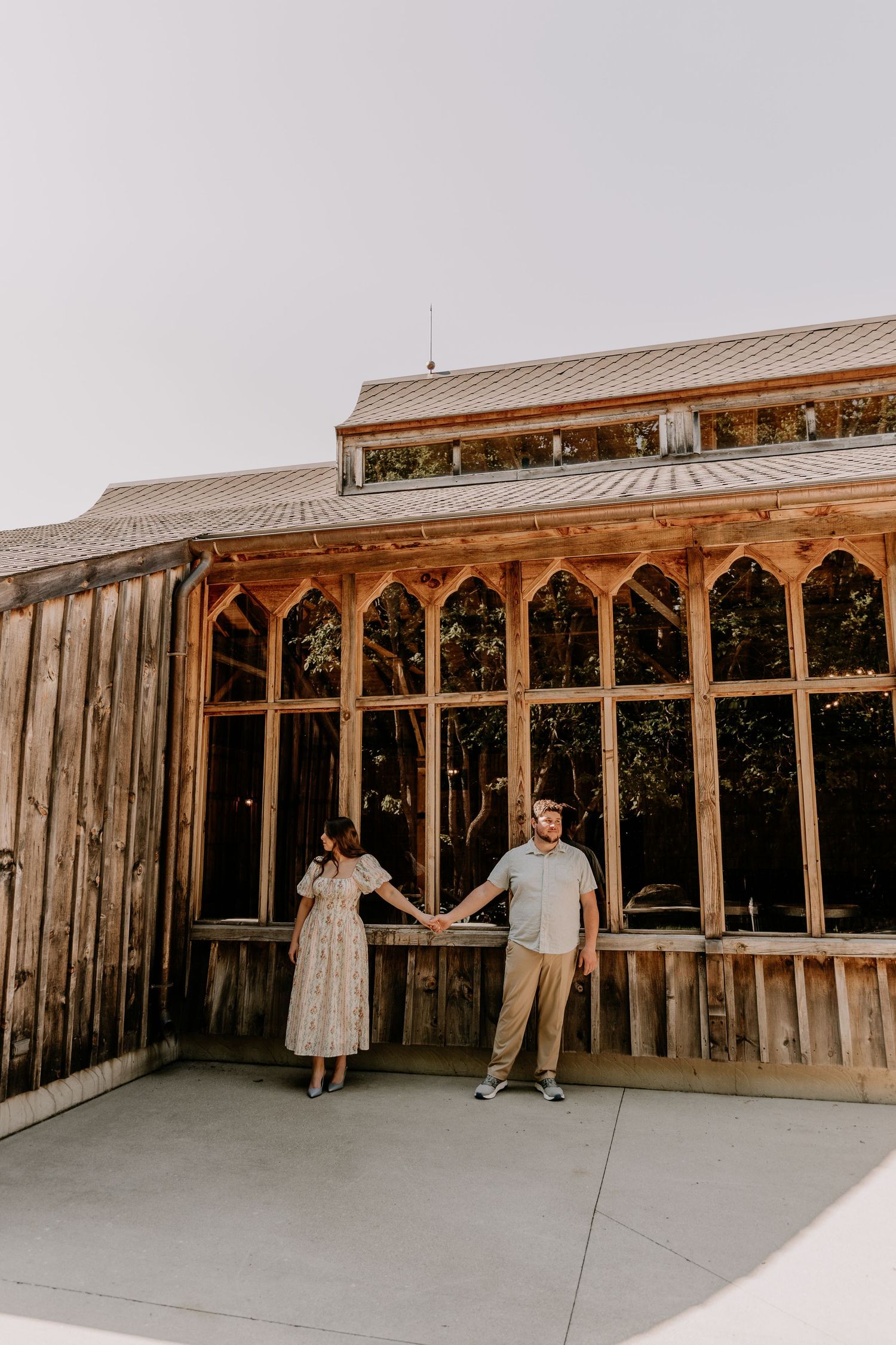 Couple holding hands in front of a rustic wooden greenhouse building with arched windows on a cloudy day.