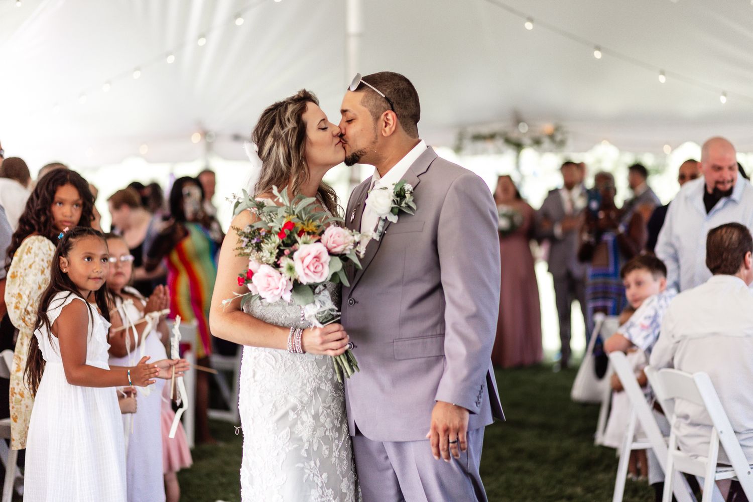 Wedding ceremony under white tent with guests seated on folding chairs.