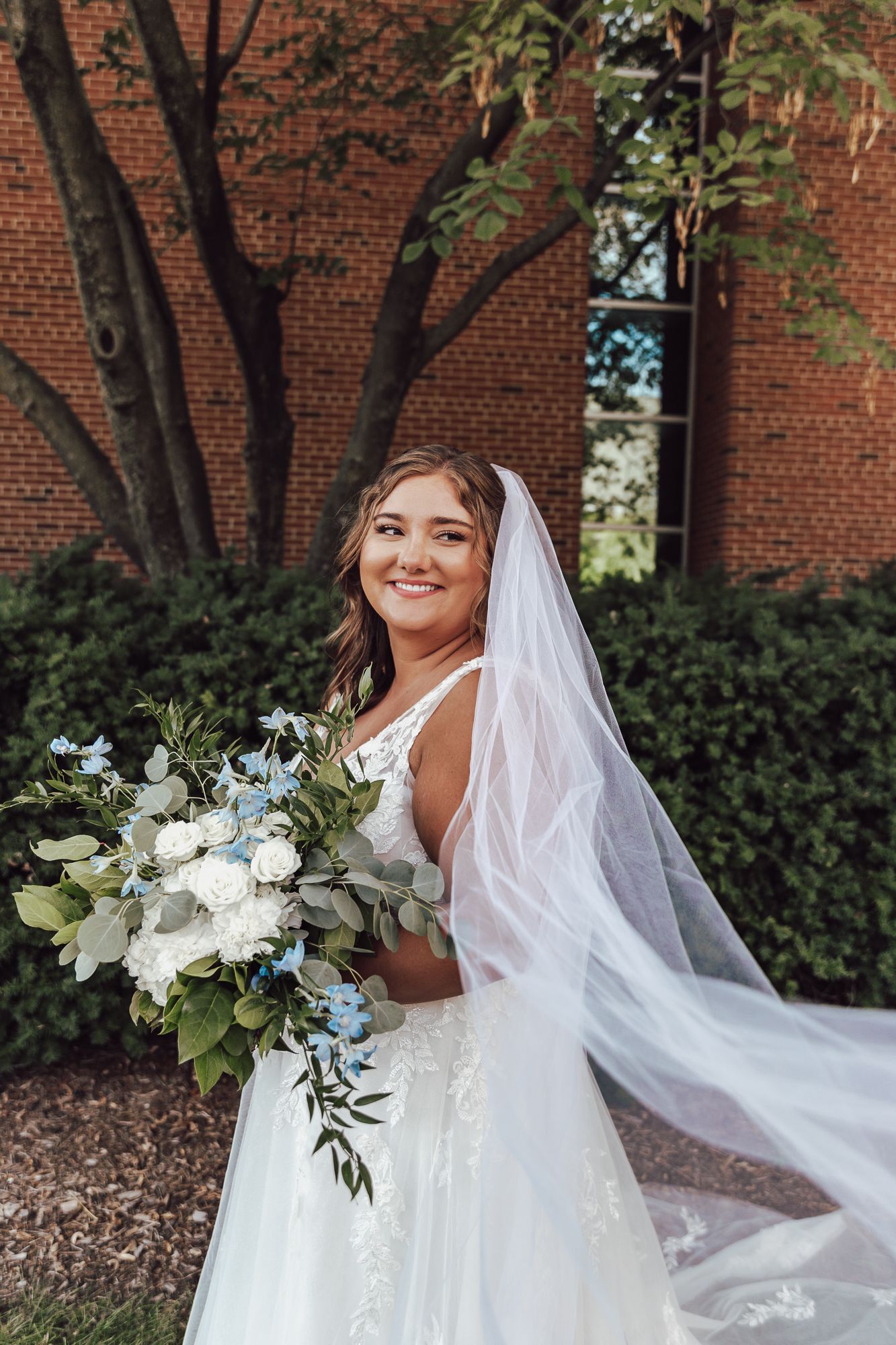 Radiant bride in white gown holds elegant bouquet while posing next to brick building.