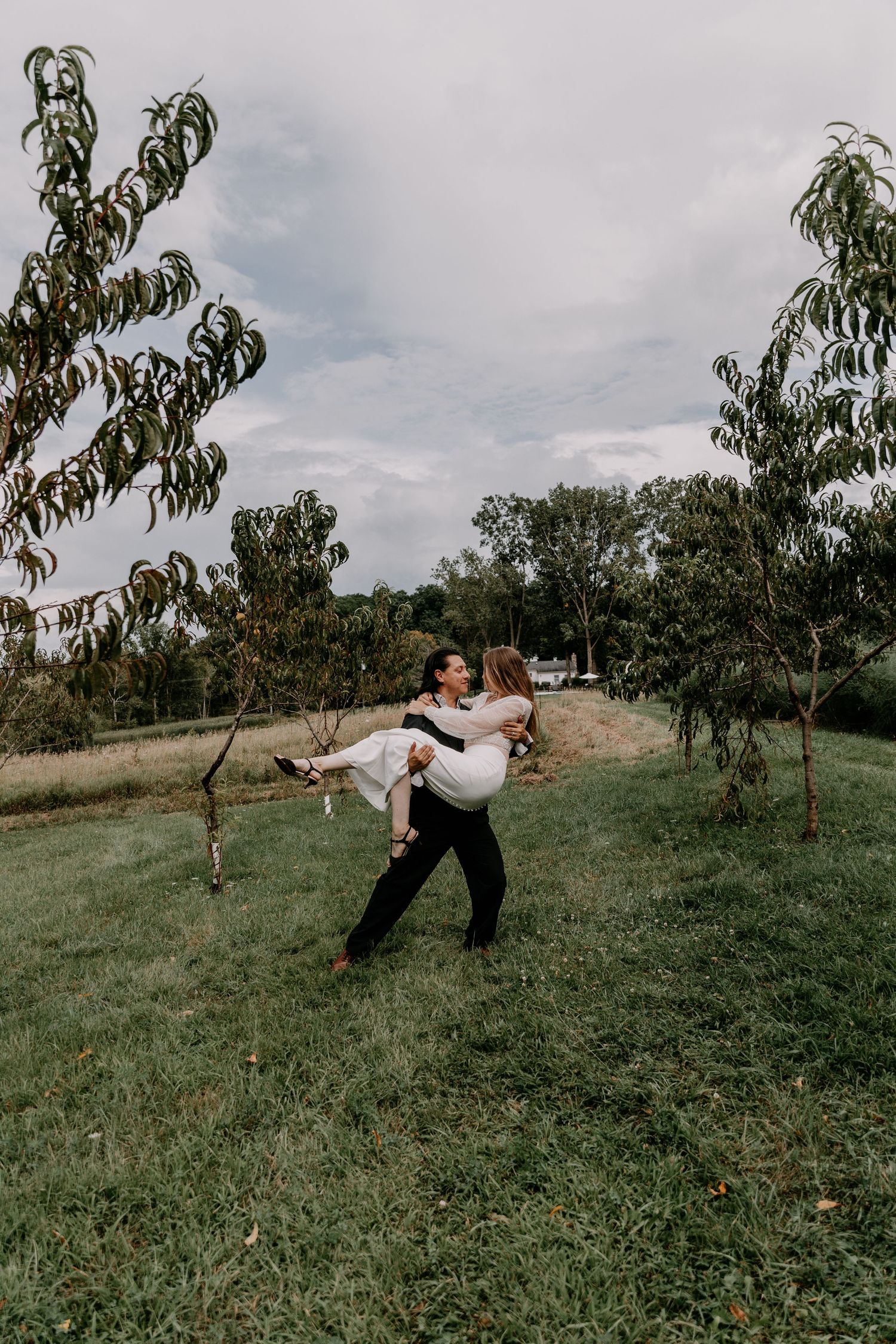Groom carrying bride in white dress among trees in outdoor setting.
