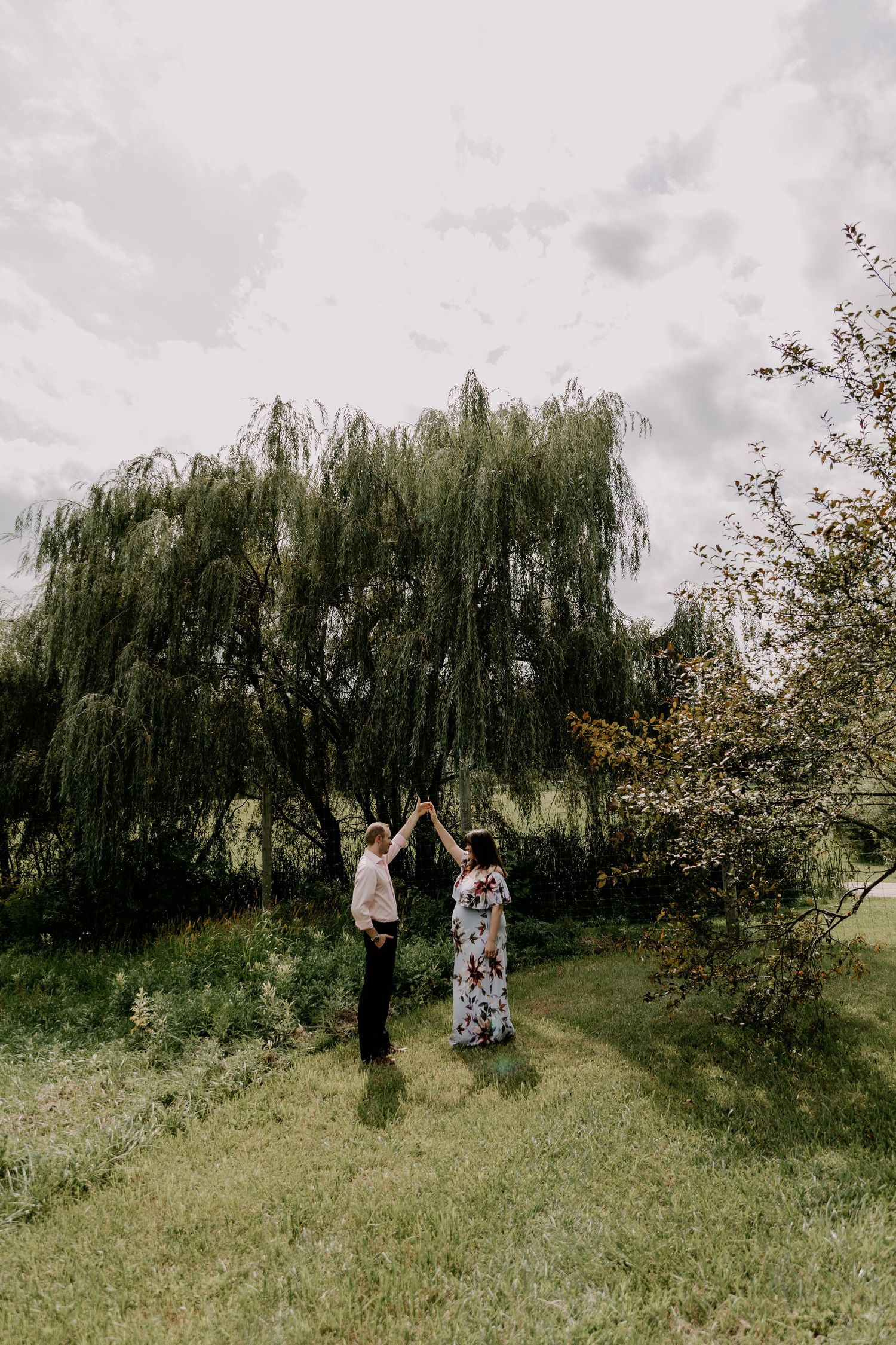 Two people dance and twirl joyfully on a grassy lawn beneath majestic weeping willow trees.