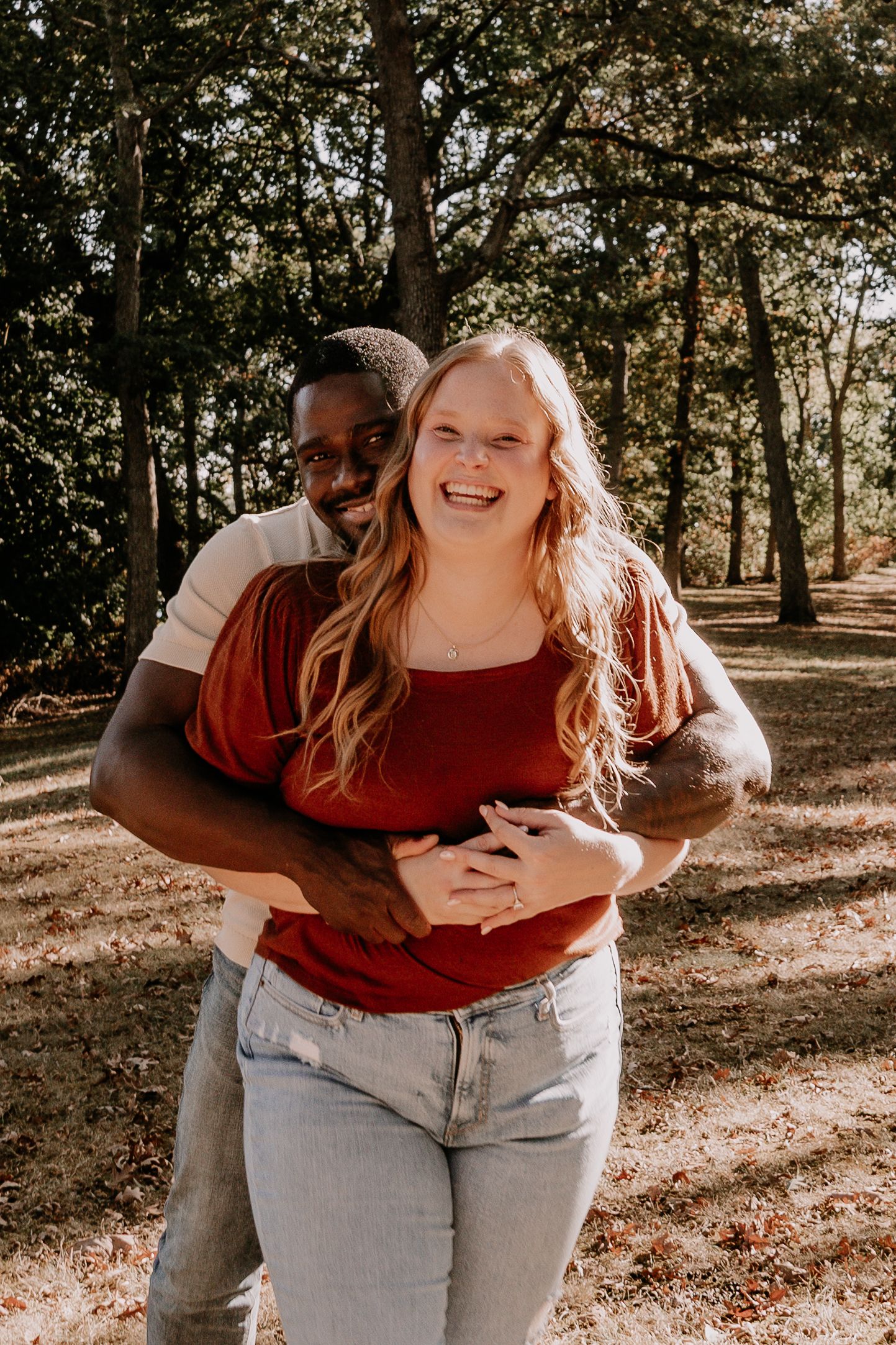 A couple embraces and laughs together during a sunny autumn photoshoot in a wooded park setting.