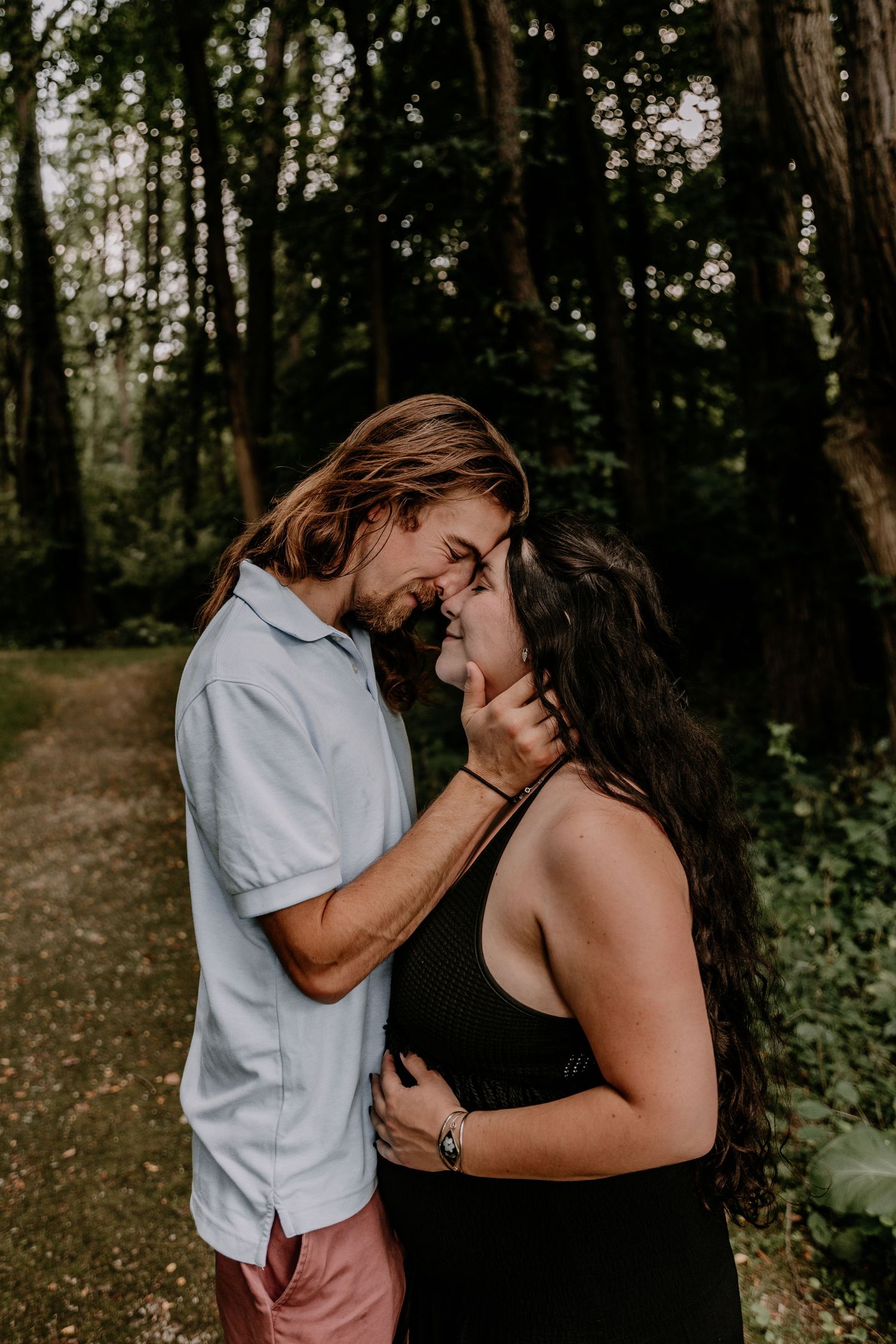 A couple shares a romantic forehead touch on a forest path, the man in a light shirt and the woman in a black dress.