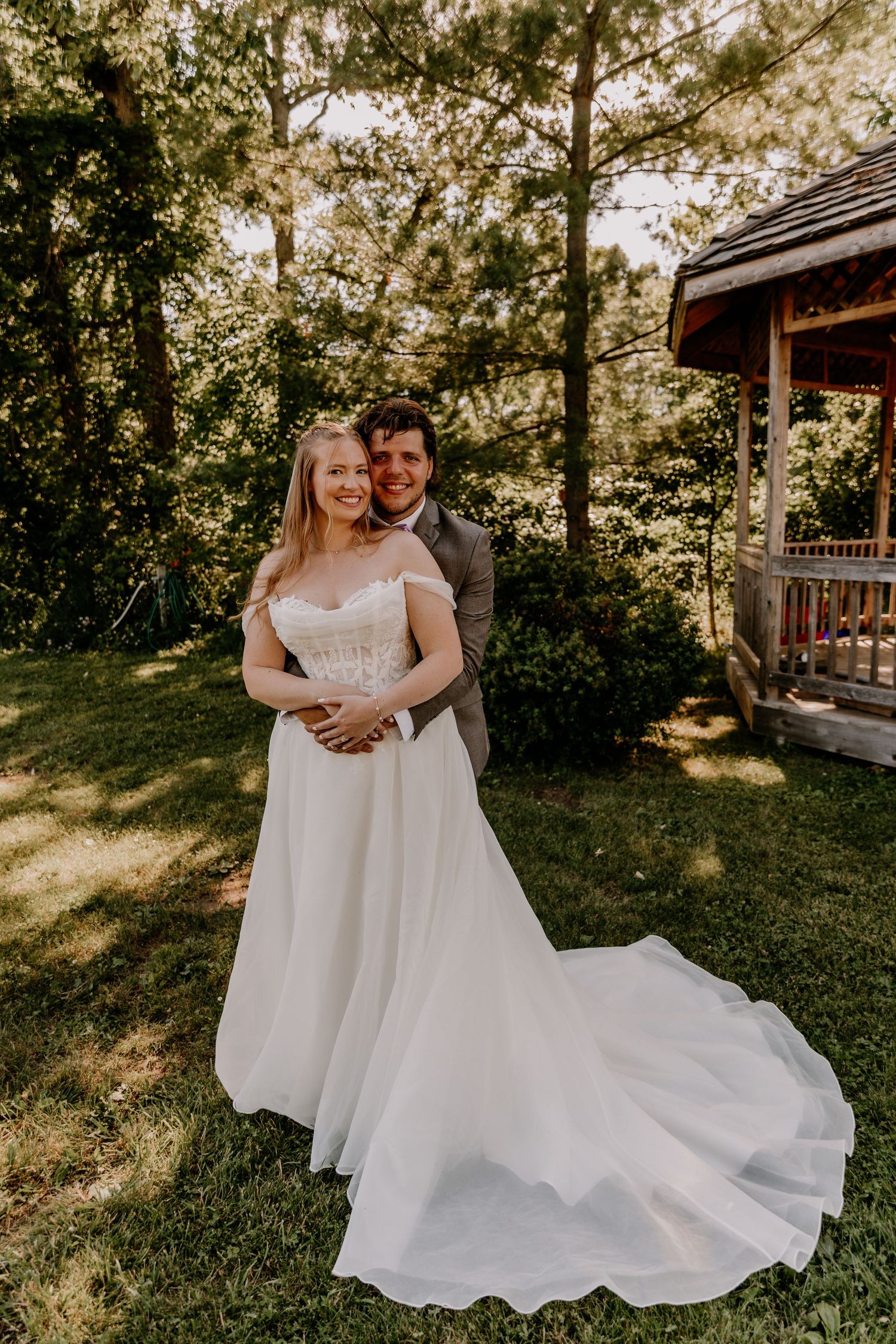 Bride in white off-shoulder gown with train stands with groom in gray suit outdoors near gazebo.