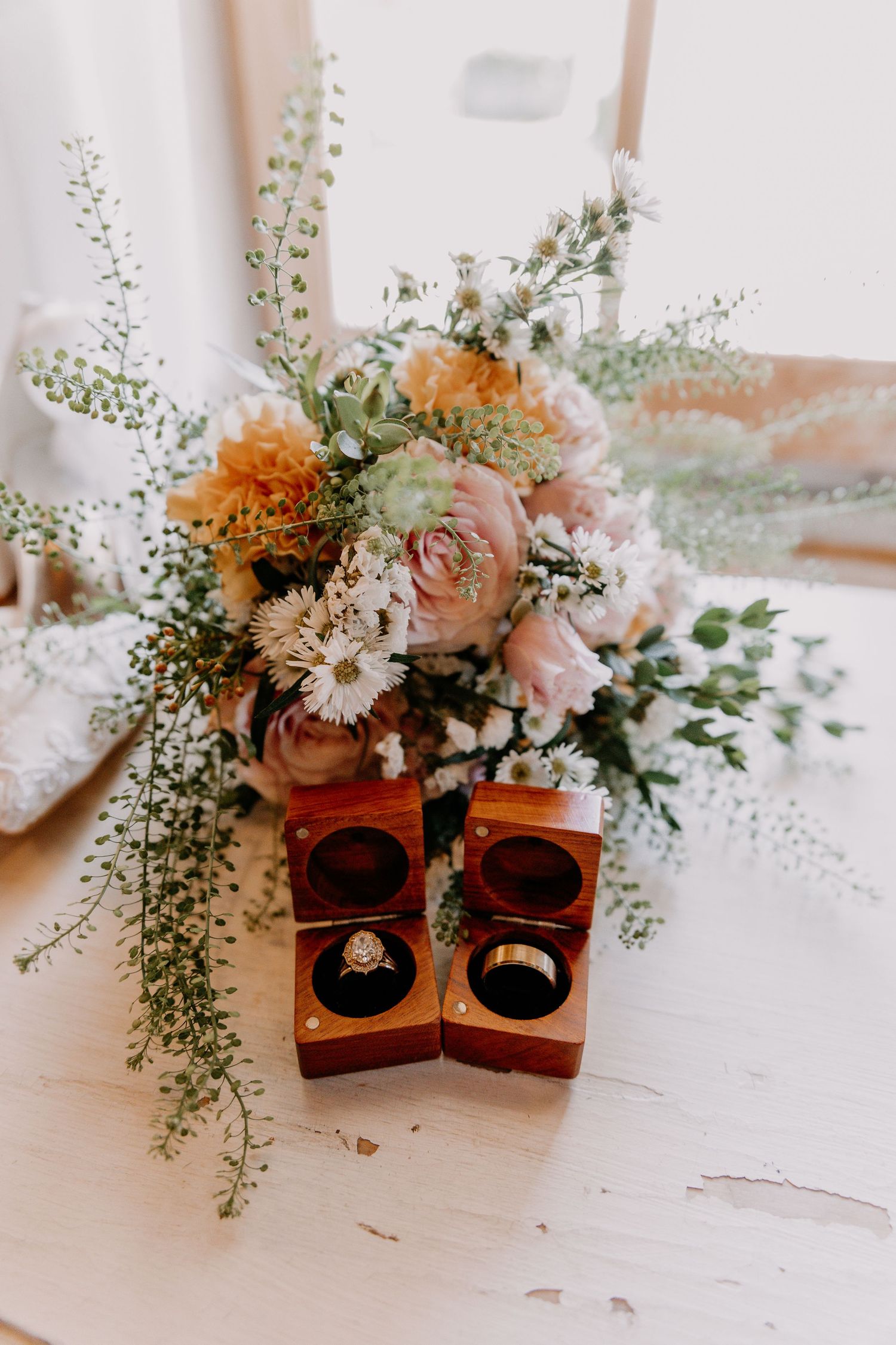 Wedding rings in wooden boxes displayed in front of a romantic bouquet of blush pink, orange, and white flowers.