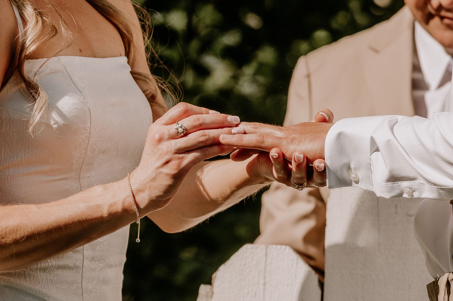 Bride placing a wedding ring on groom's finger during an outdoor ceremony in natural sunlight.