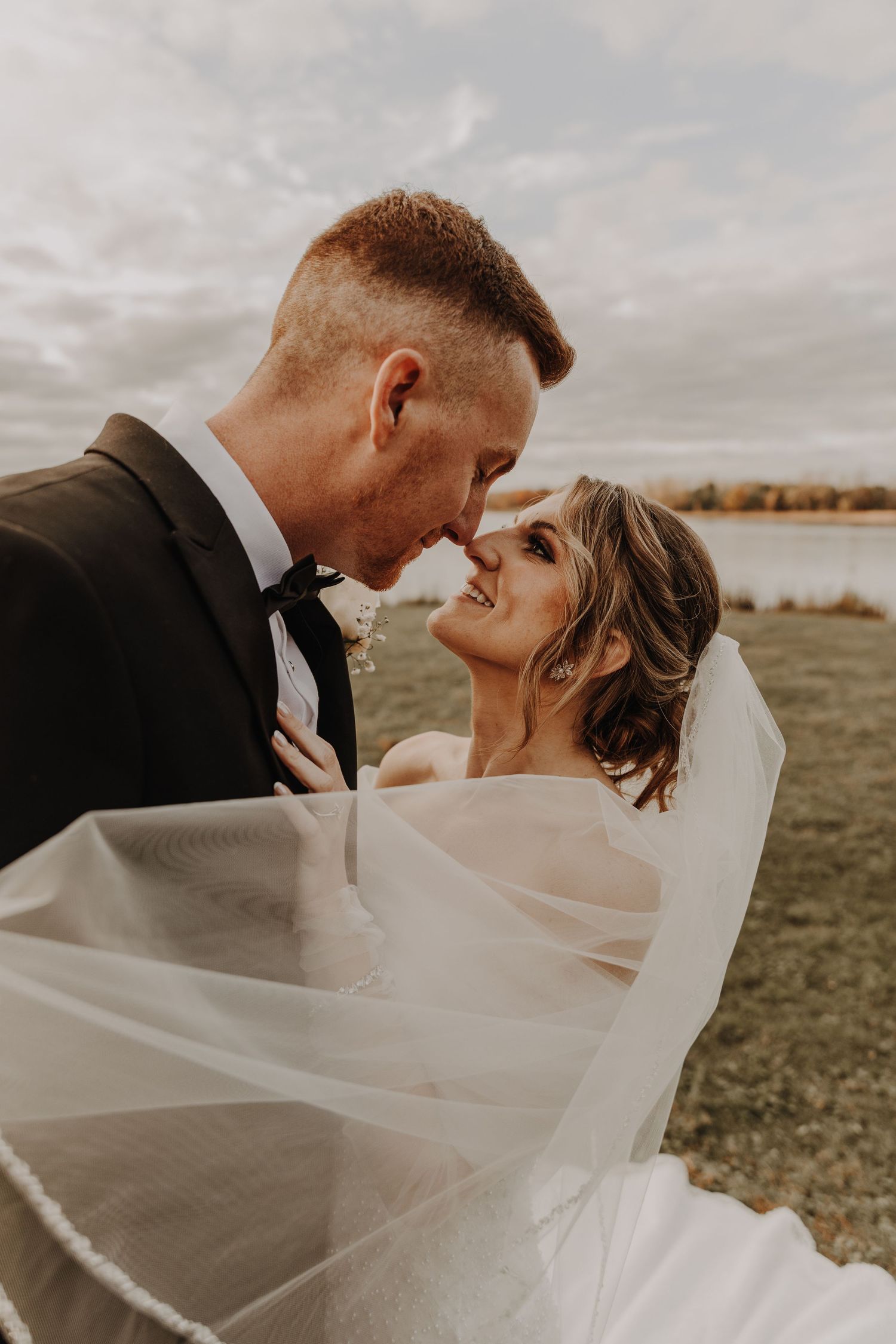A romantic wedding couple embraces outdoors as the bride's veil flows in the wind during their autumn photoshoot.