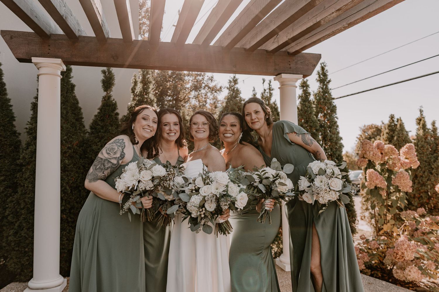 Wedding party wearing sage green dresses poses together on a white porch during sunset.