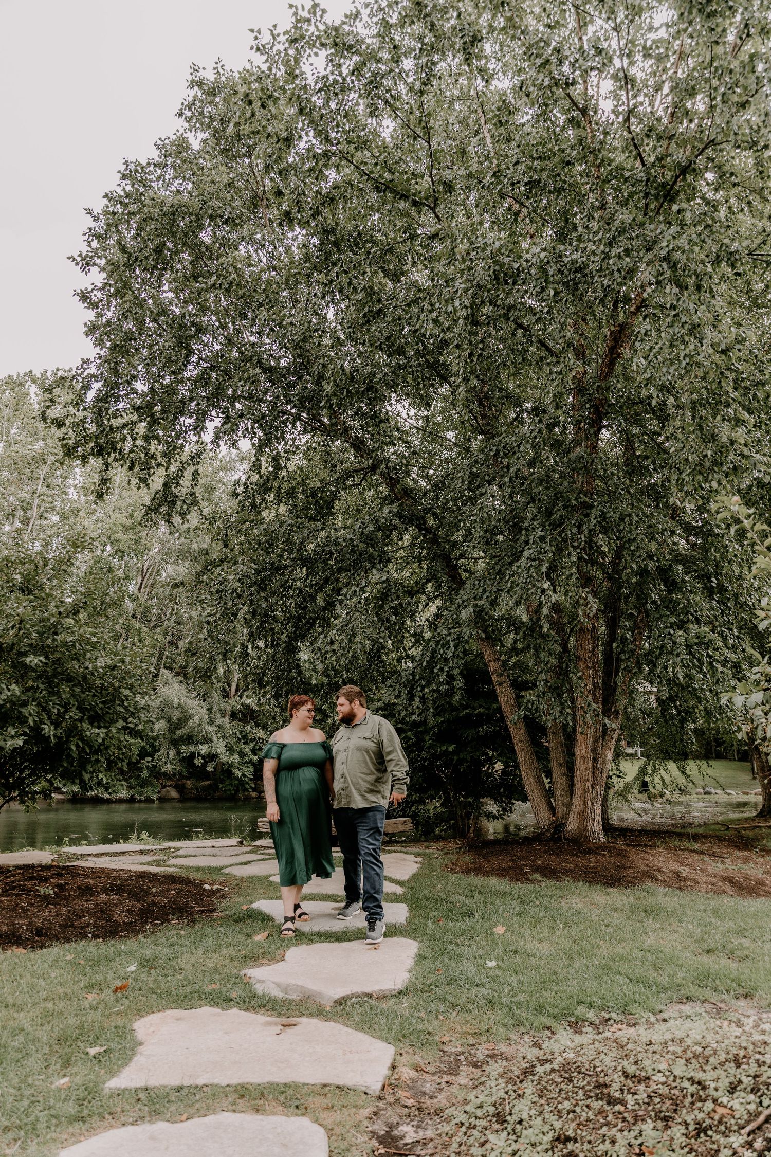 A couple walks hand-in-hand along a stone path in a lush green park near a pond.