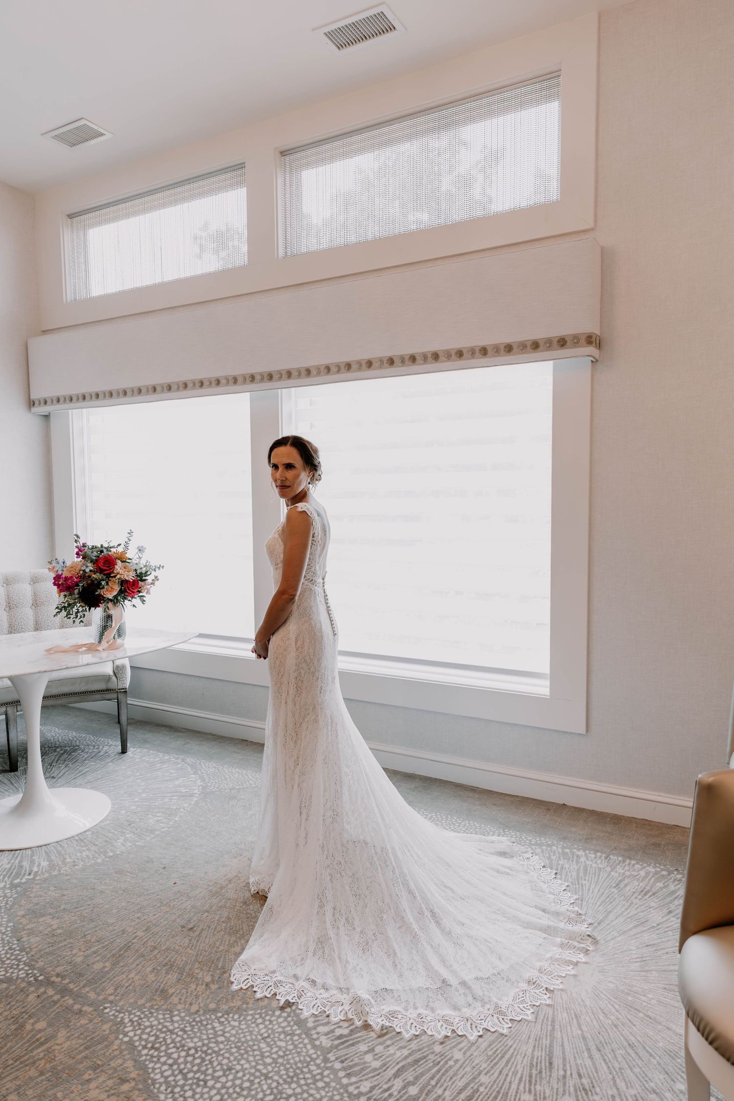Elegant bride in lace wedding dress with long train poses in bright white room with large windows.