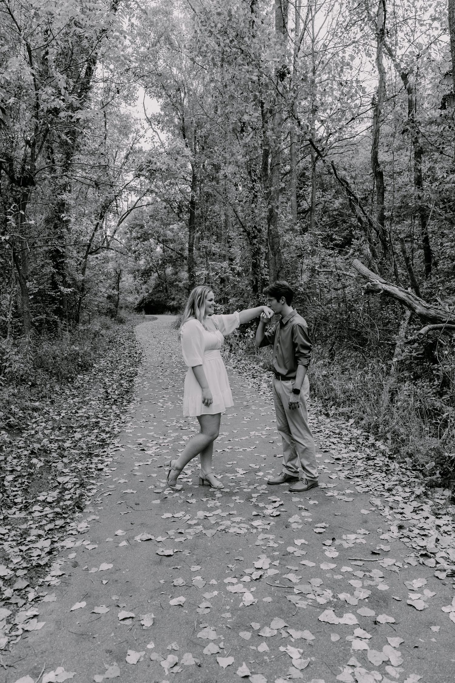 A couple dances playfully together on a leaf-covered forest path in black and white.