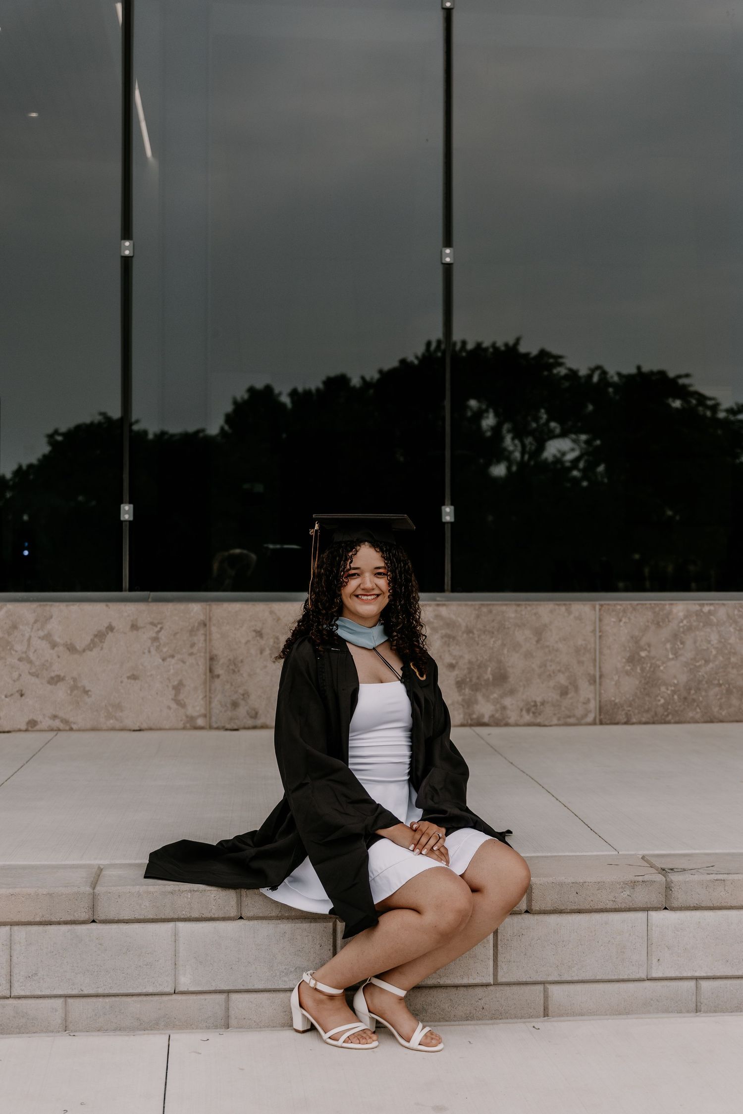 Figure seated on stone steps wearing a black coat and white dress on an overcast day.