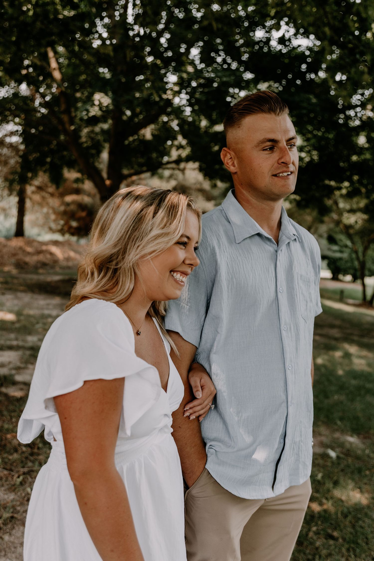 A couple shares a candid moment outdoors, wearing white and light blue casual attire in a natural park setting.