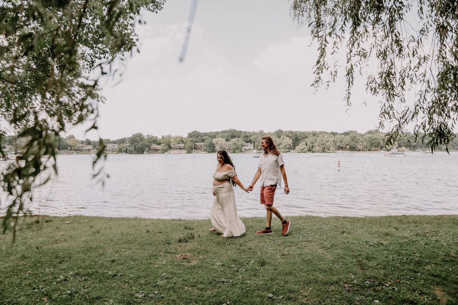 Two women holding hands and walking by a serene lakeside on a grassy shore, framed by weeping willow branches.