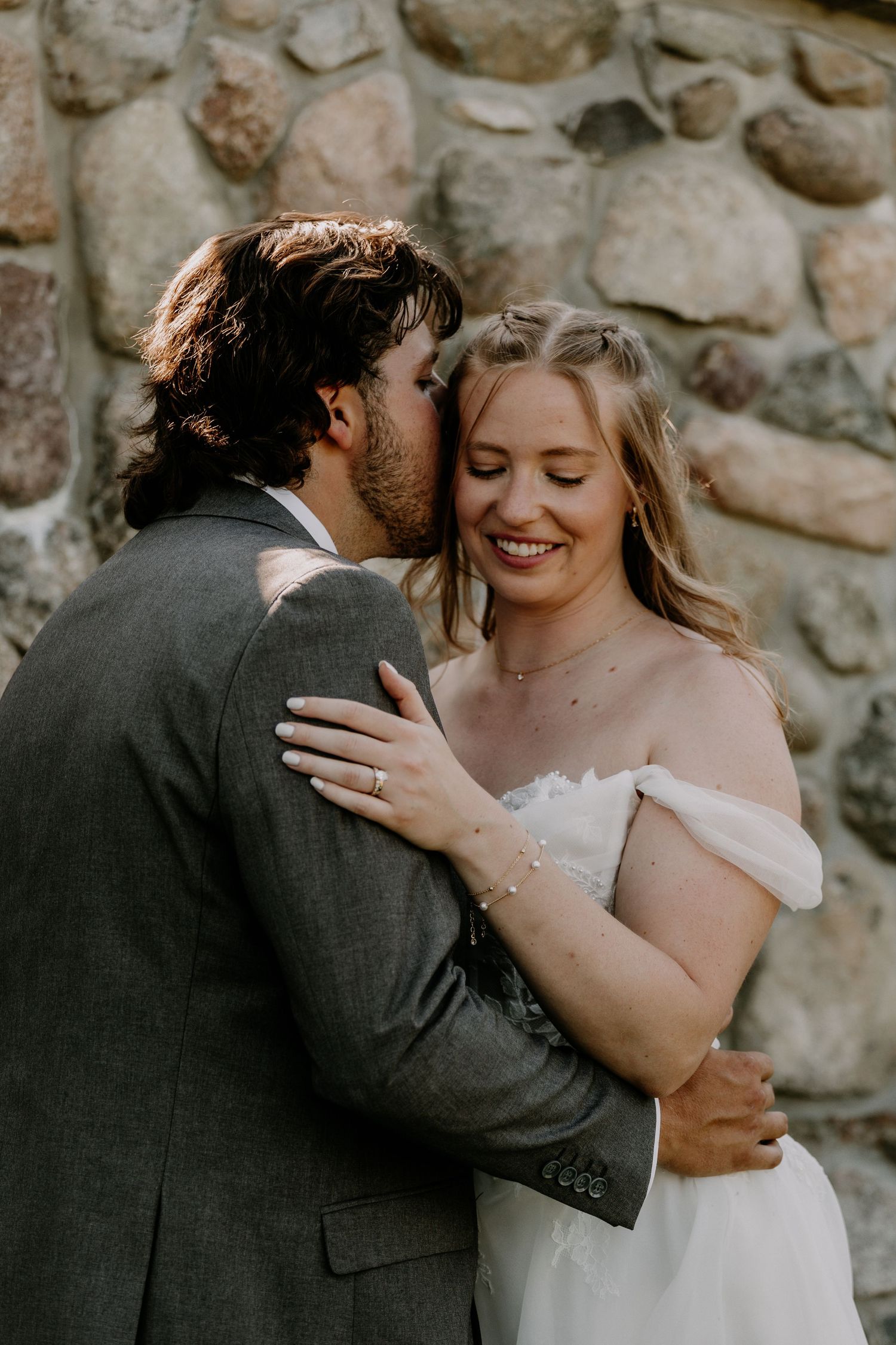 Groom in gray suit kisses bride in white off-shoulder gown as she smiles, standing before a rustic stone wall.