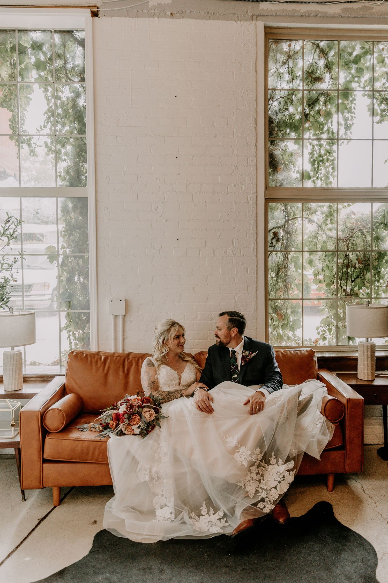 A romantic wedding couple lounges together on a brown leather couch in a bright, modern space with large windows.