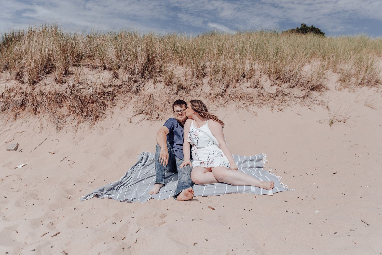 A couple sits together on a blanket among coastal sand dunes on a bright afternoon.