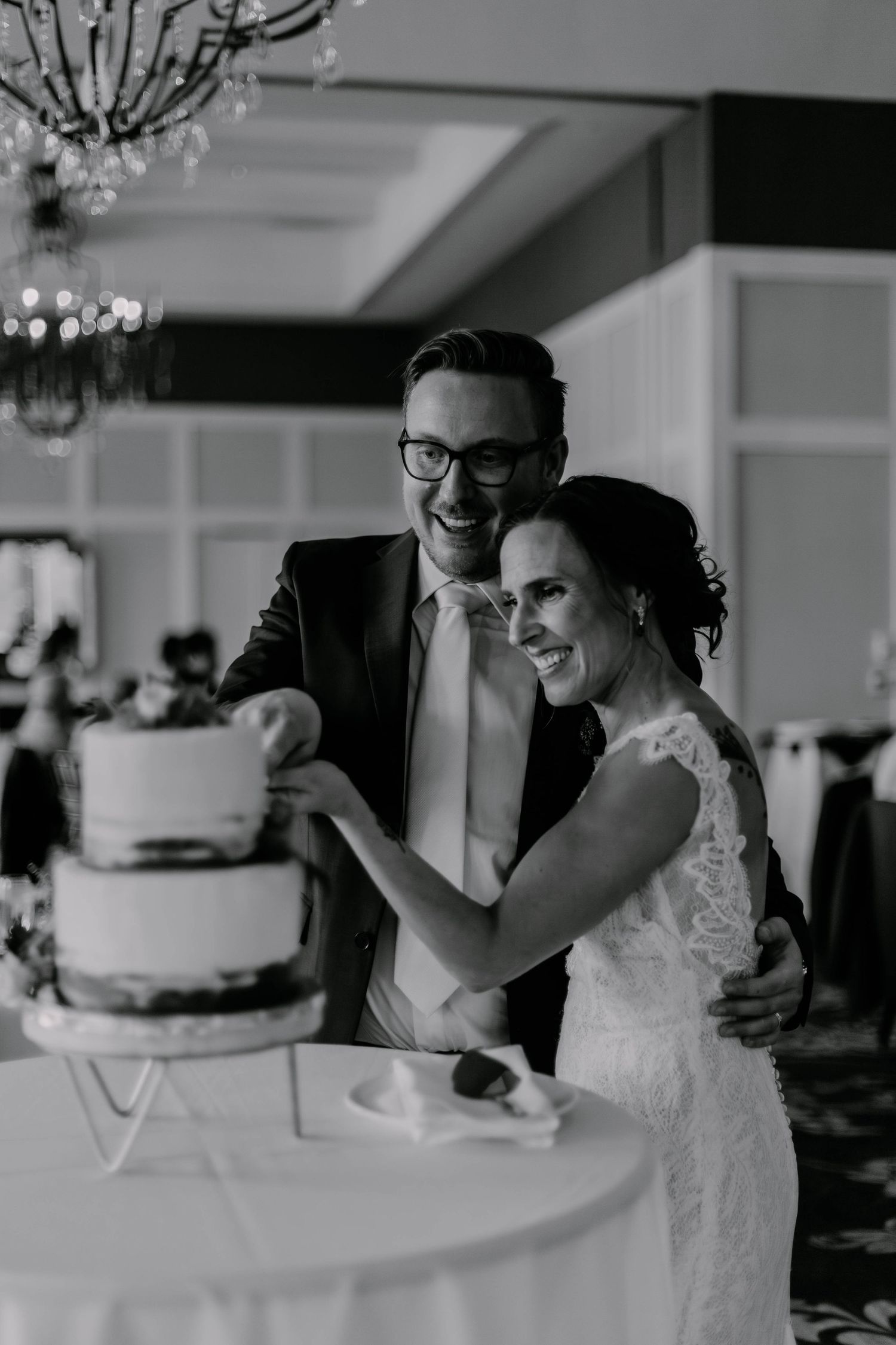 A black and white photo of a couple joyfully cutting their wedding cake together under elegant chandeliers.