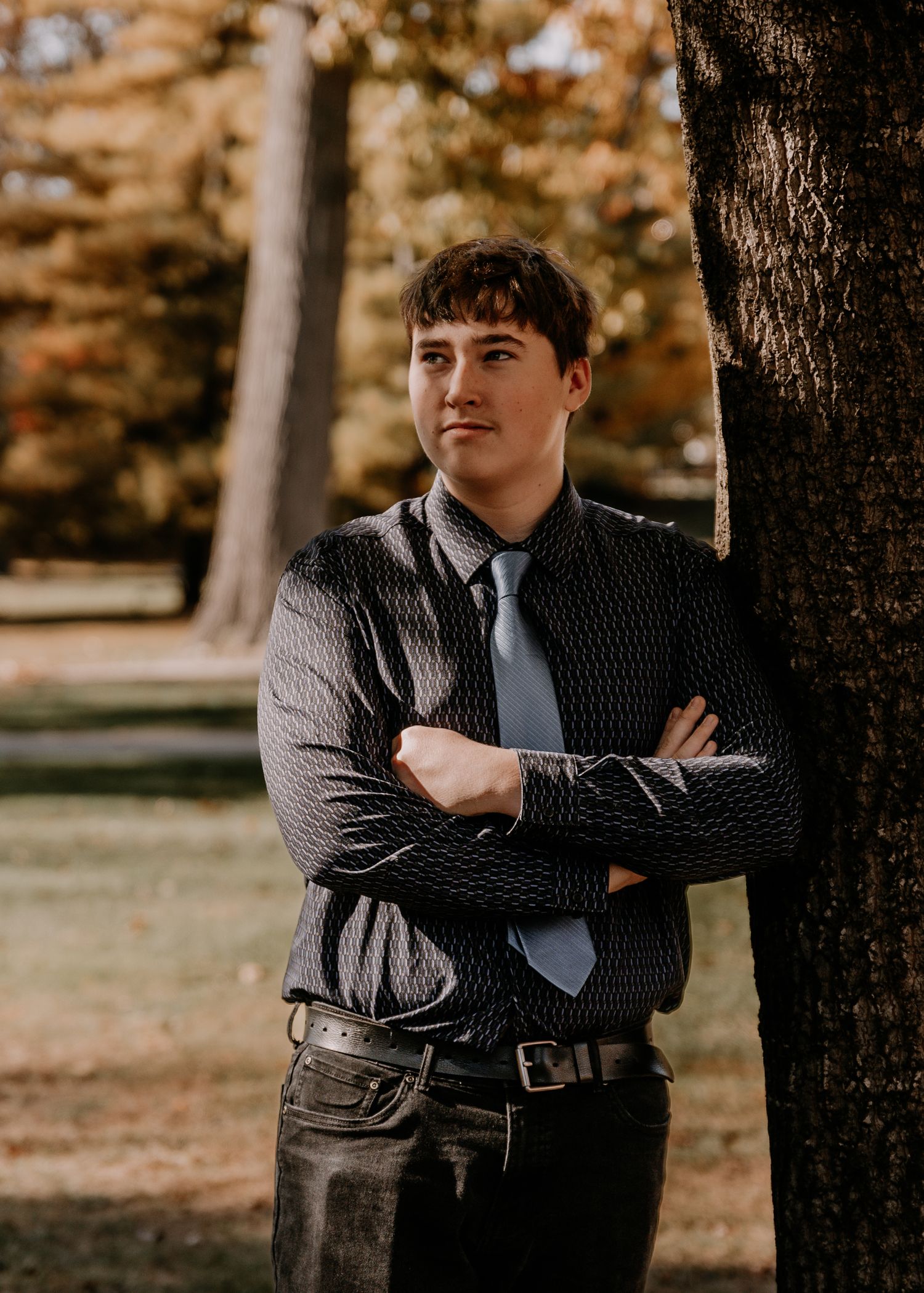 Person in dark shirt and tie poses confidently against tree in autumn setting.