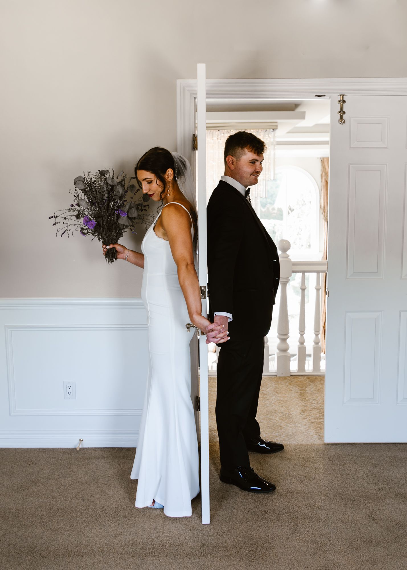Couple shares first look moment behind door with bride holding purple bouquet.