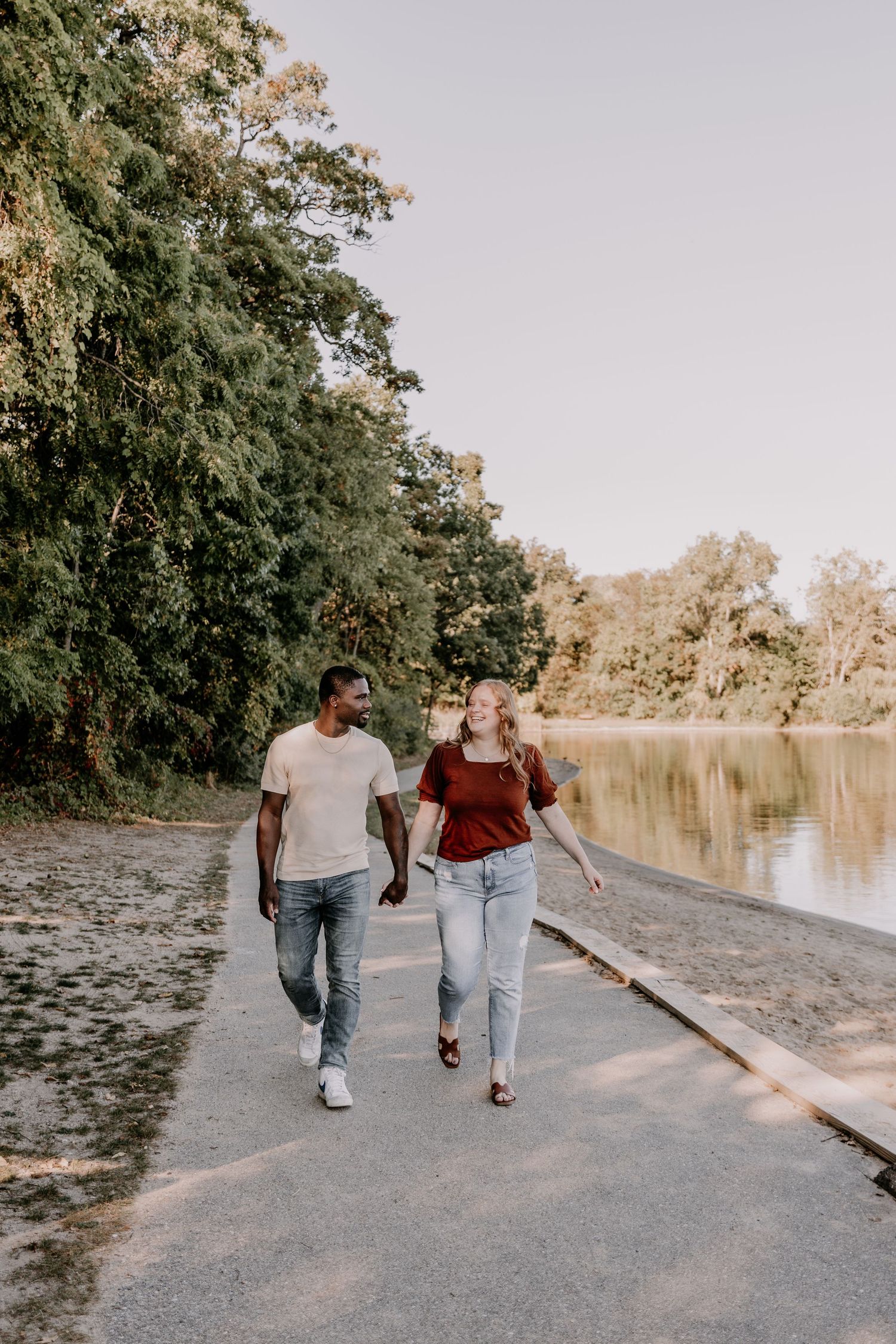 A couple walks together on a peaceful path beside a serene lake surrounded by lush trees on an autumn day.
