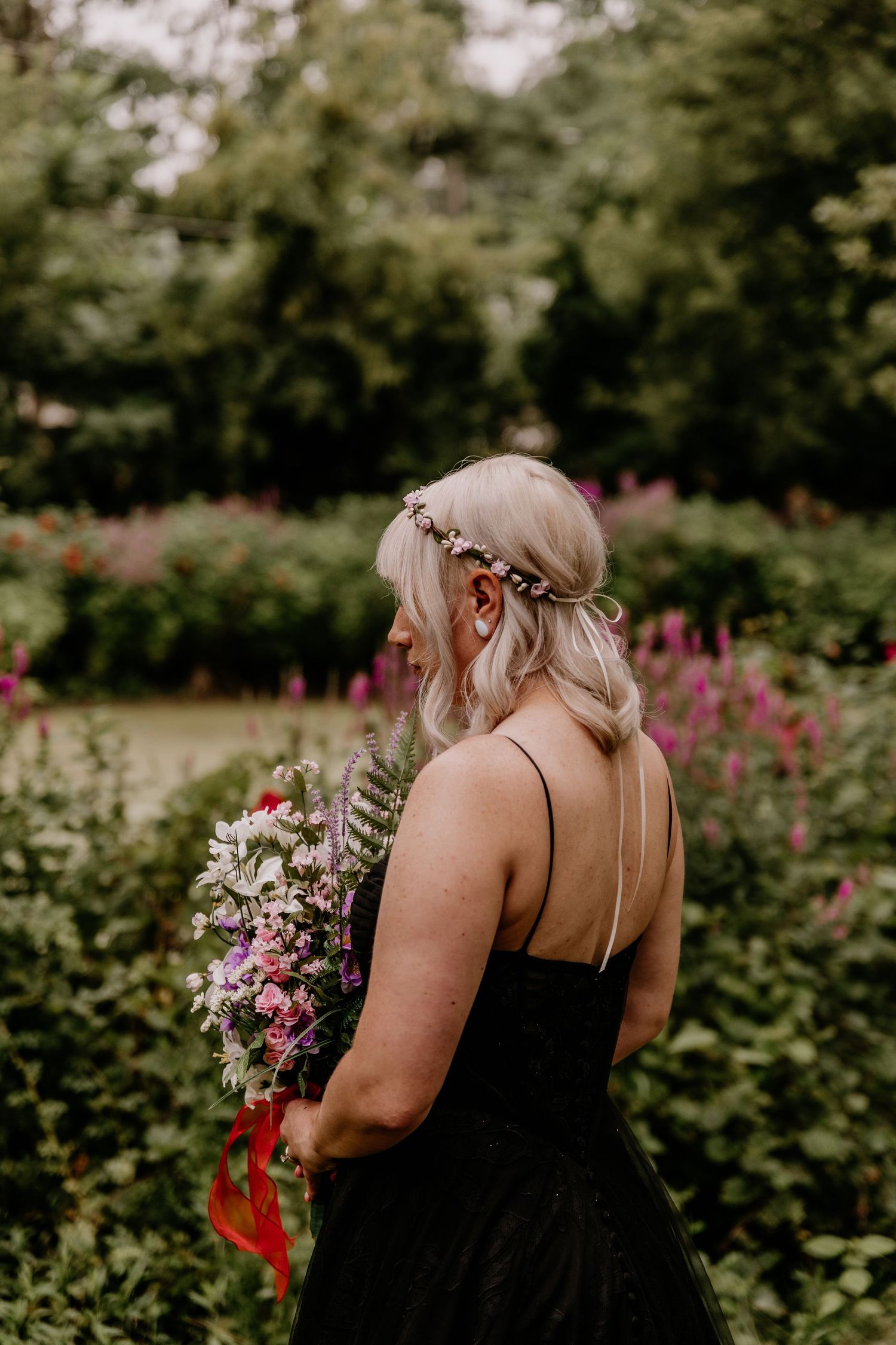 Blonde woman in black dress with floral crown holds bouquet in lush garden setting.