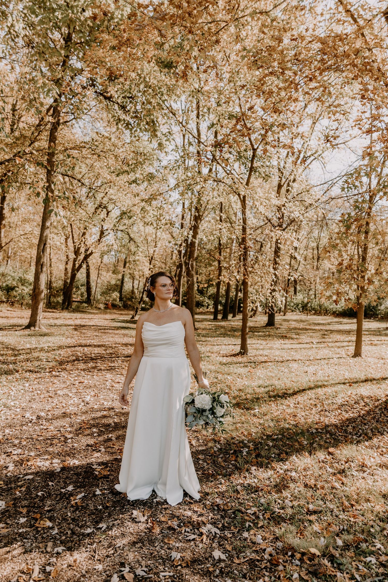 A person in a white wedding dress stands among autumn trees with fallen leaves scattered on the ground in a park setting.