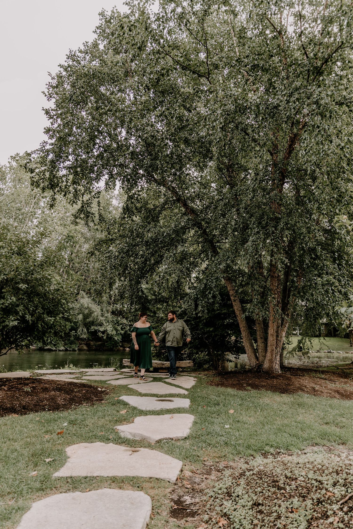 Couple holding hands walking on stepping stones through a lush green park with large trees near a pond.