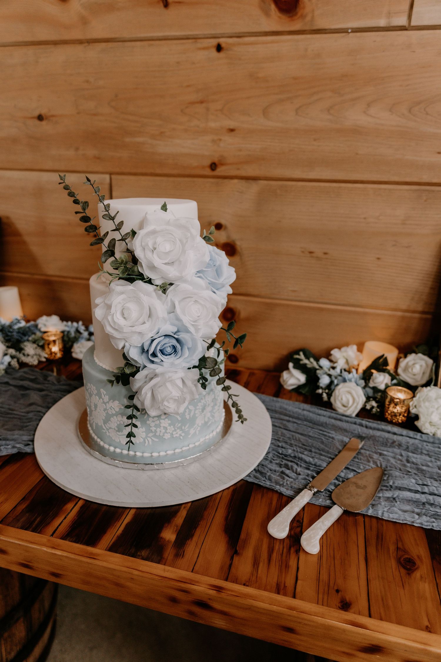 Elegant white rose wedding cake with blue accents and greenery on wooden table.