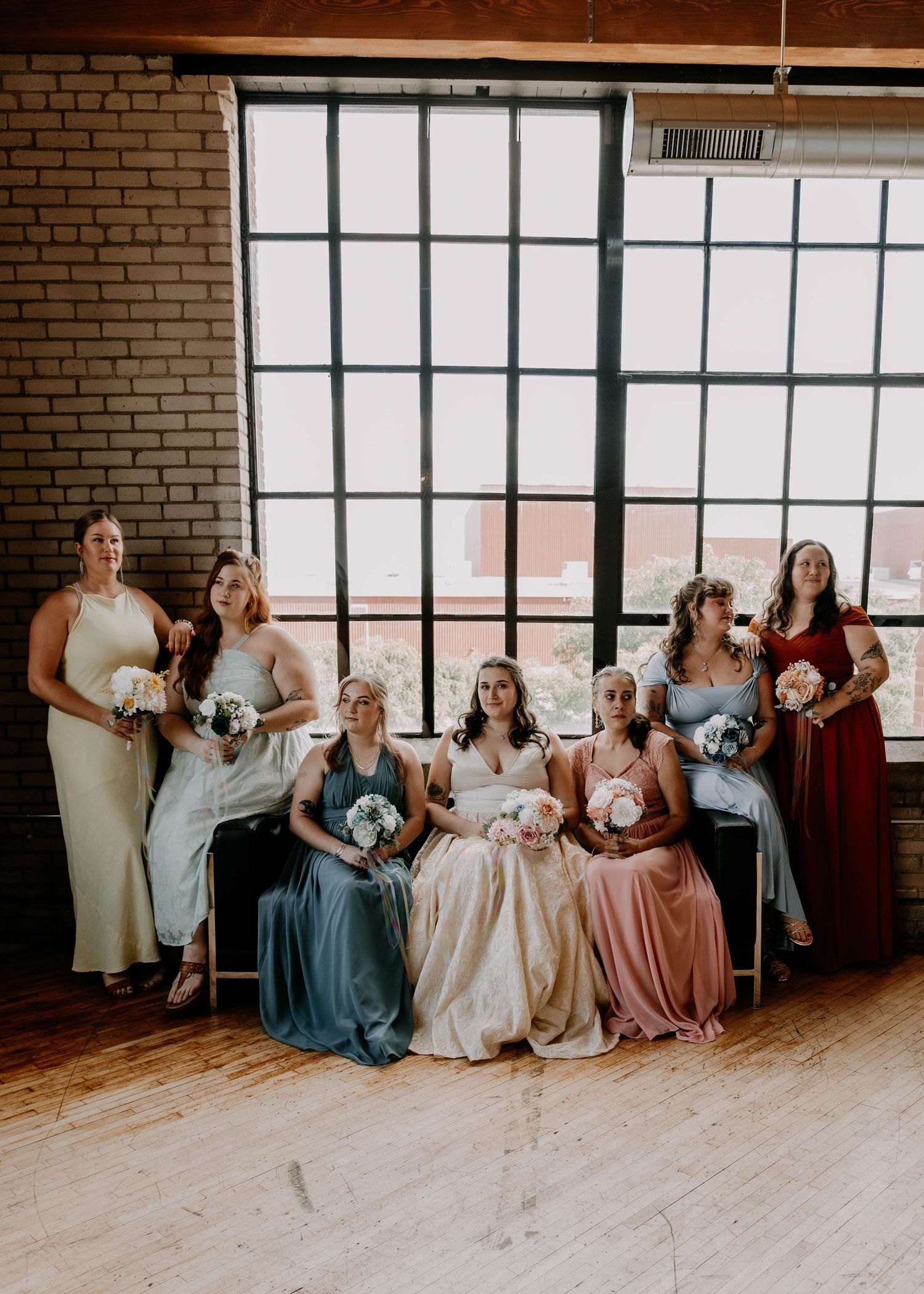 Bridal party in pastel and burgundy dresses poses by large windows in industrial brick venue.