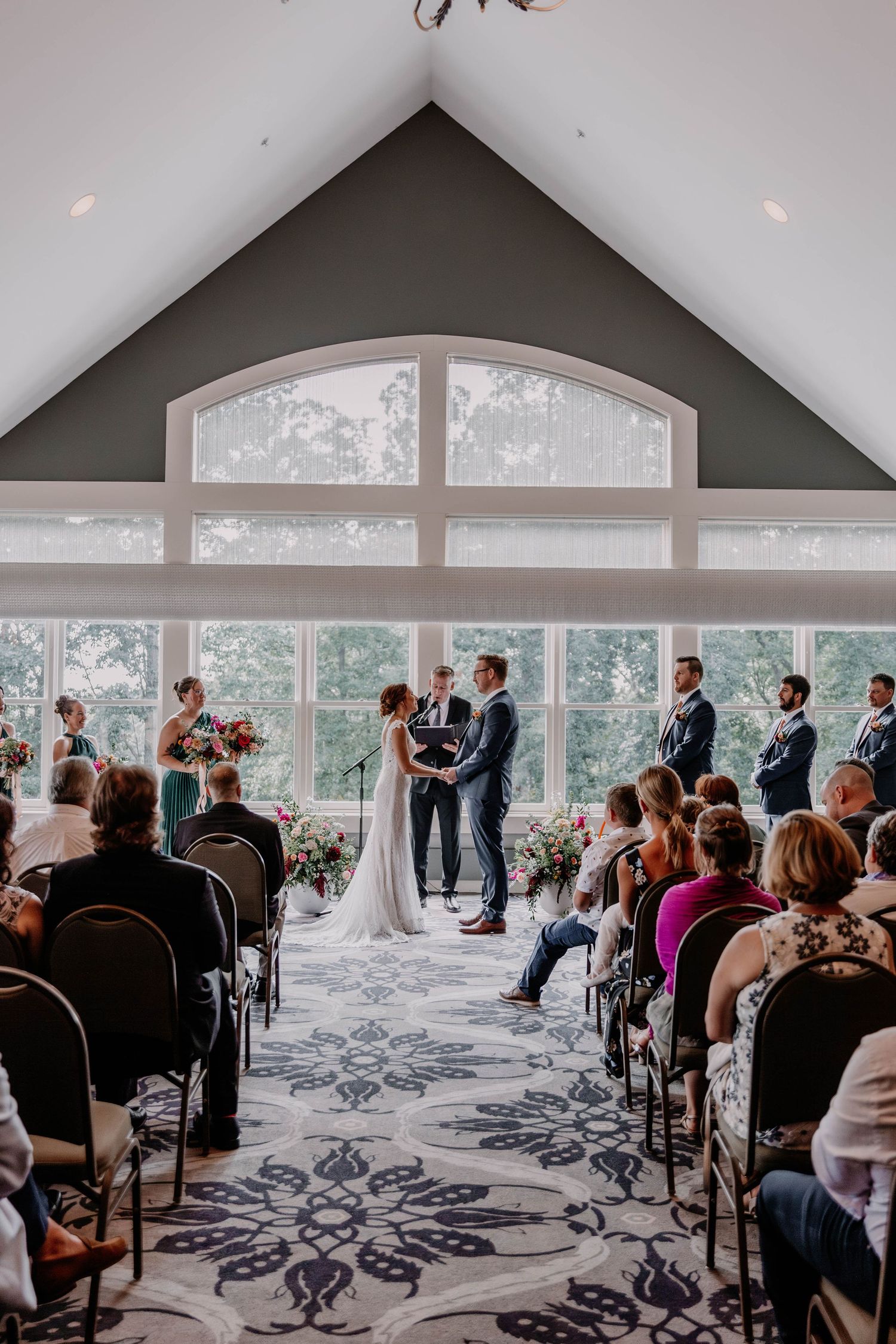 Wedding ceremony in elegant room with vaulted ceiling, large windows and ornate carpet.