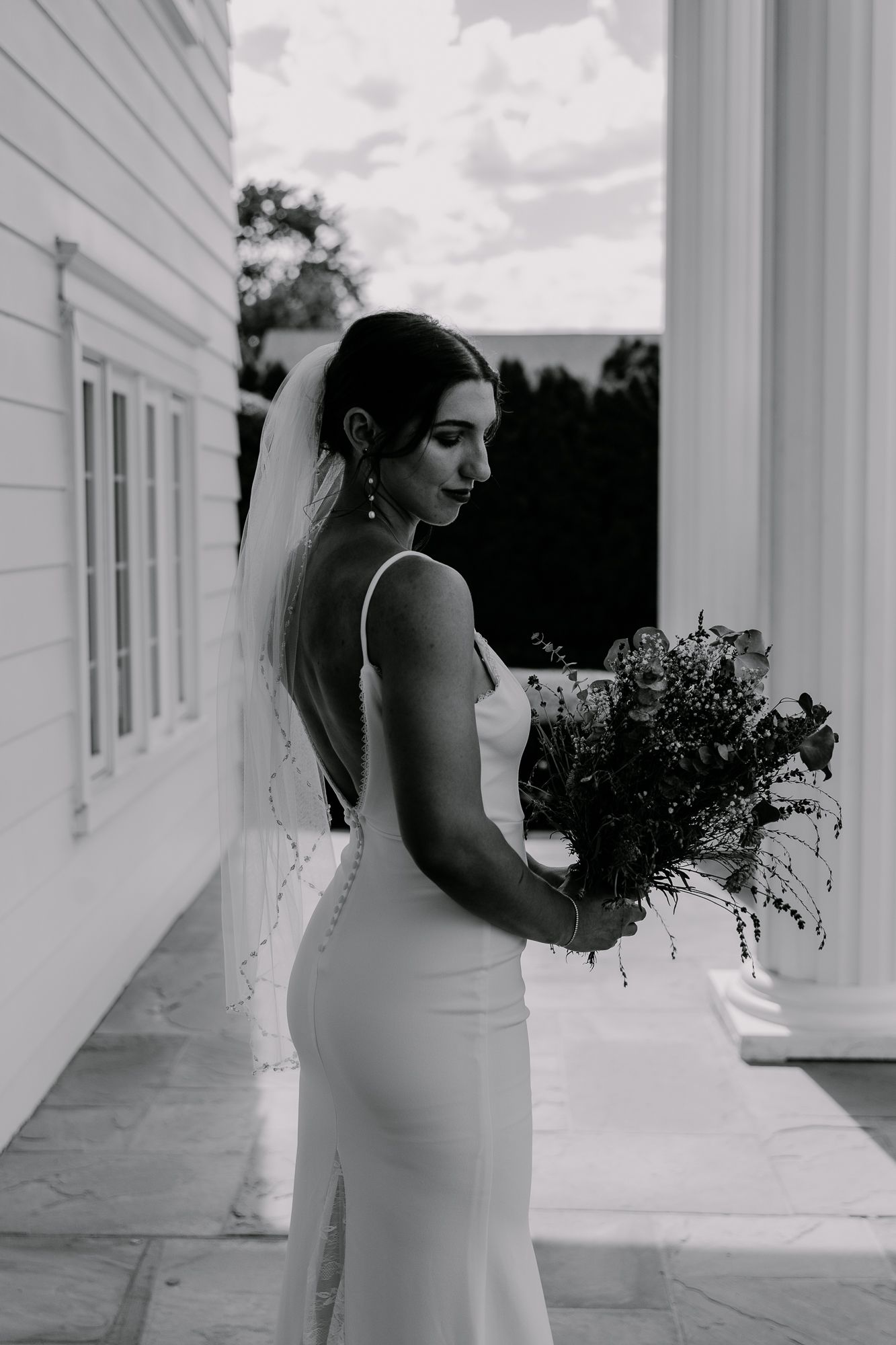 A black and white portrait of a bride holding a bouquet while standing between white columns.