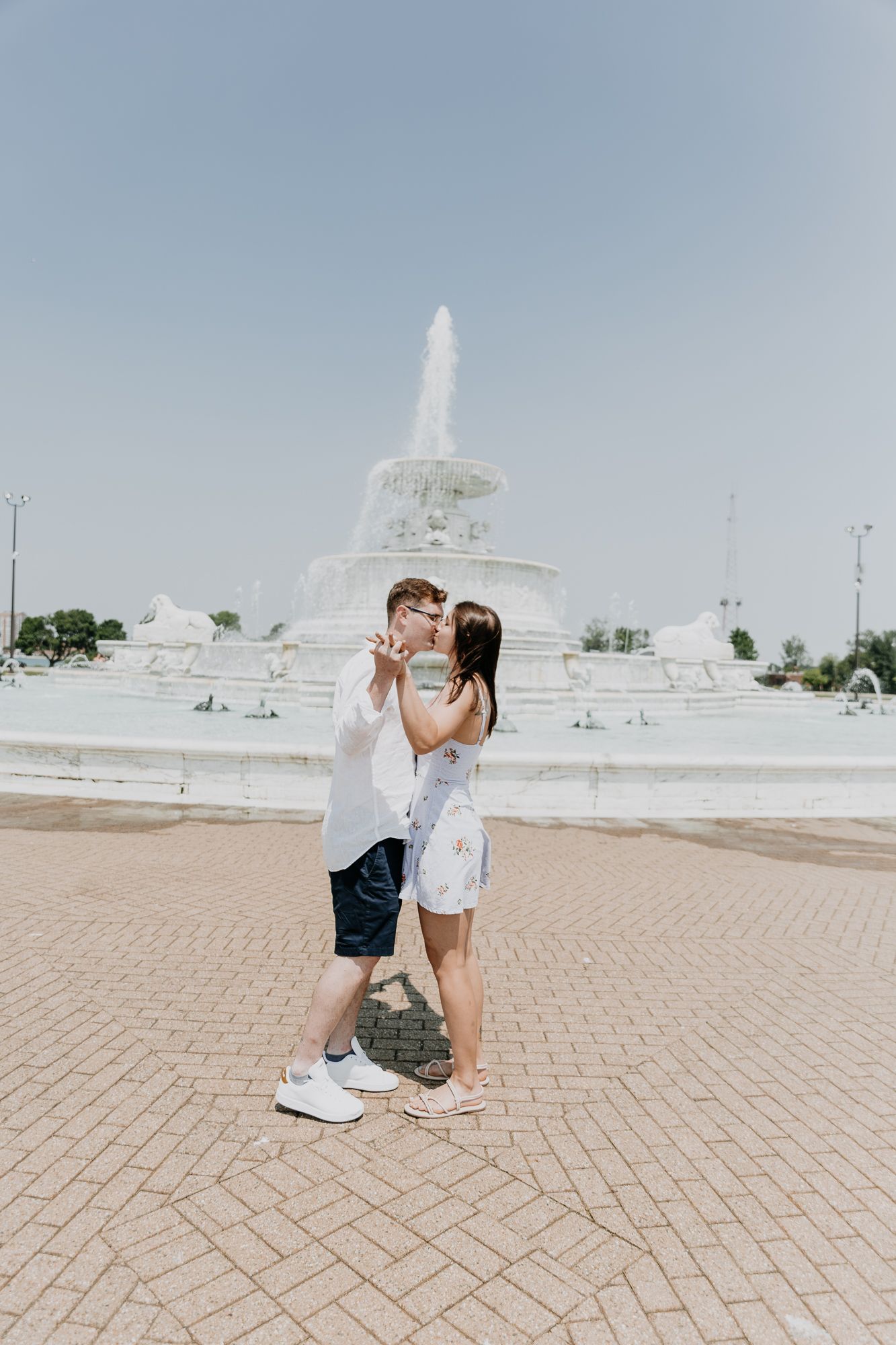 A couple embracing in front of a white fountain on a sunny day.