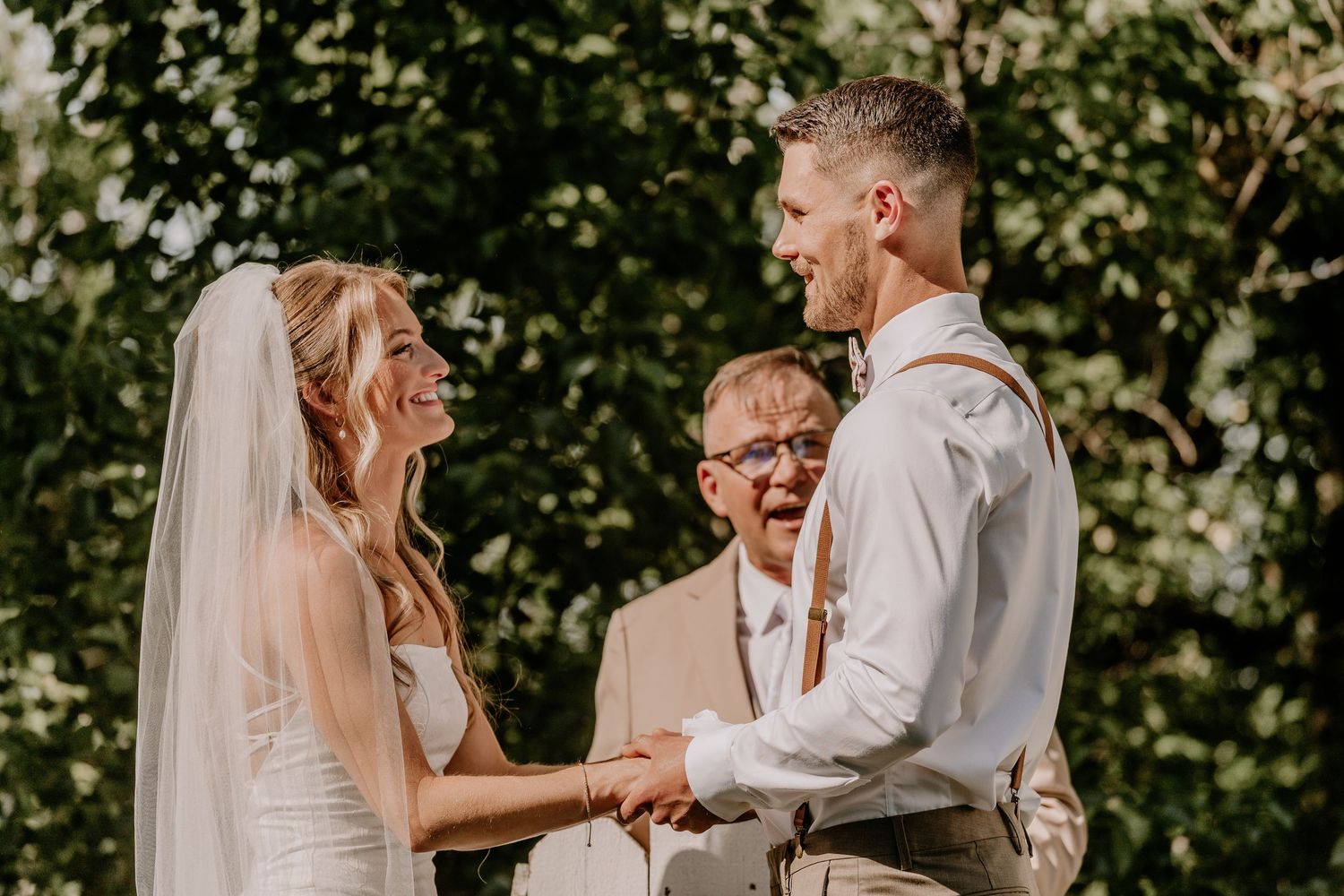 Joyful bride and groom holding hands during outdoor wedding ceremony with officiant smiling in background.
