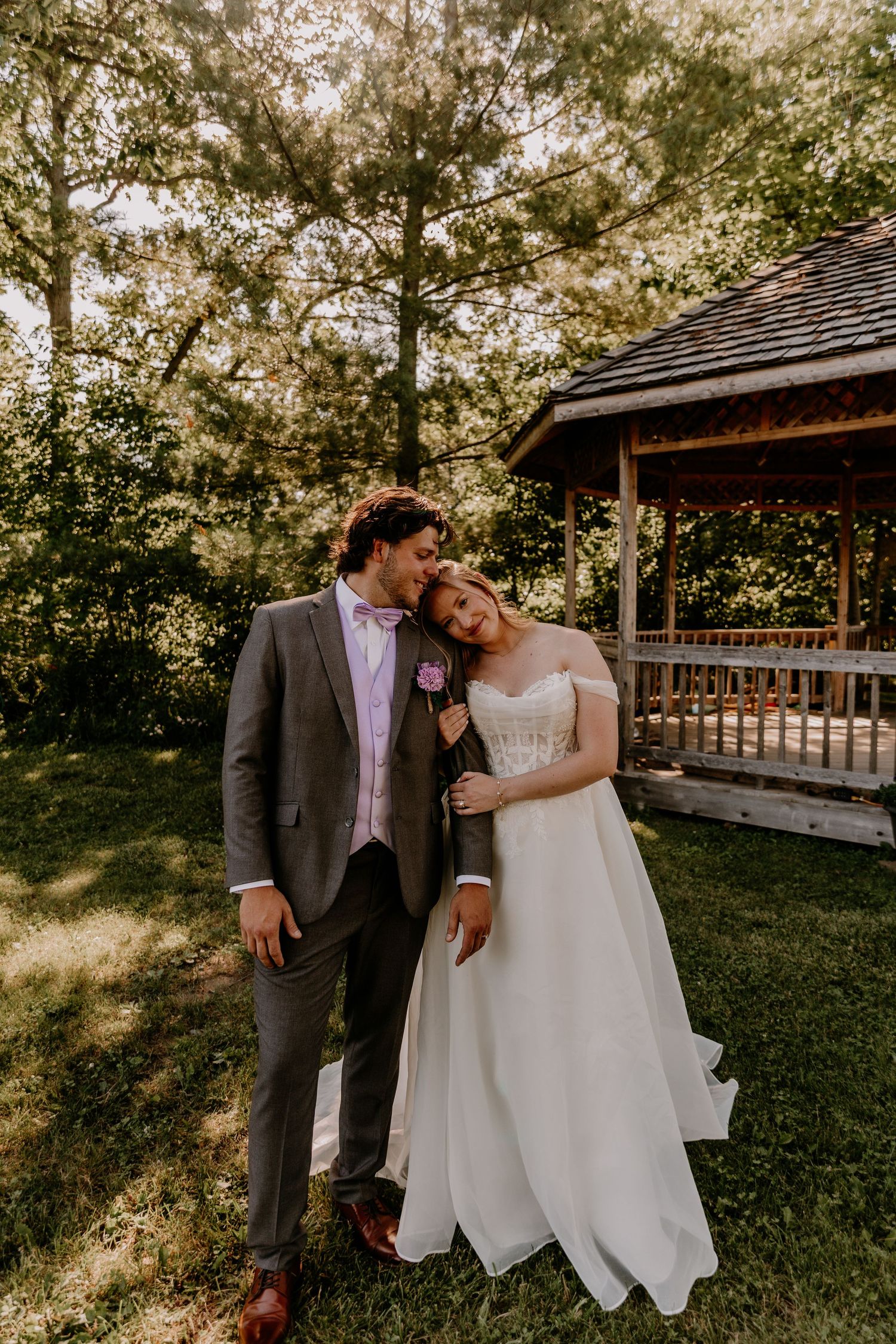 A bride and groom share a tender moment outdoors near a wooden gazebo, surrounded by lush green trees on their wedding day.