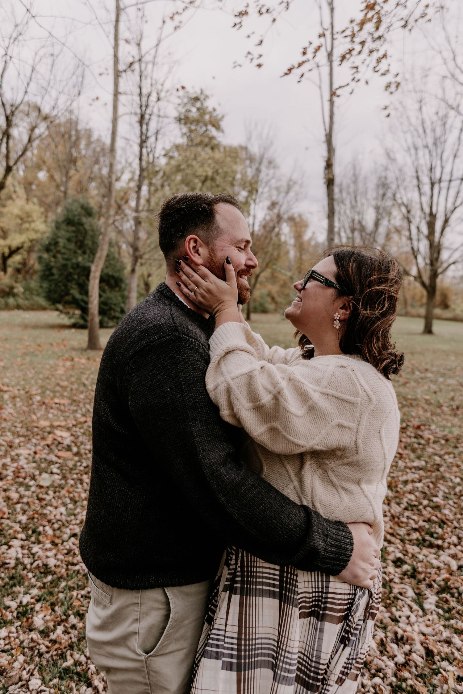 A couple shares a tender moment while embracing in a park filled with autumn leaves on the ground.