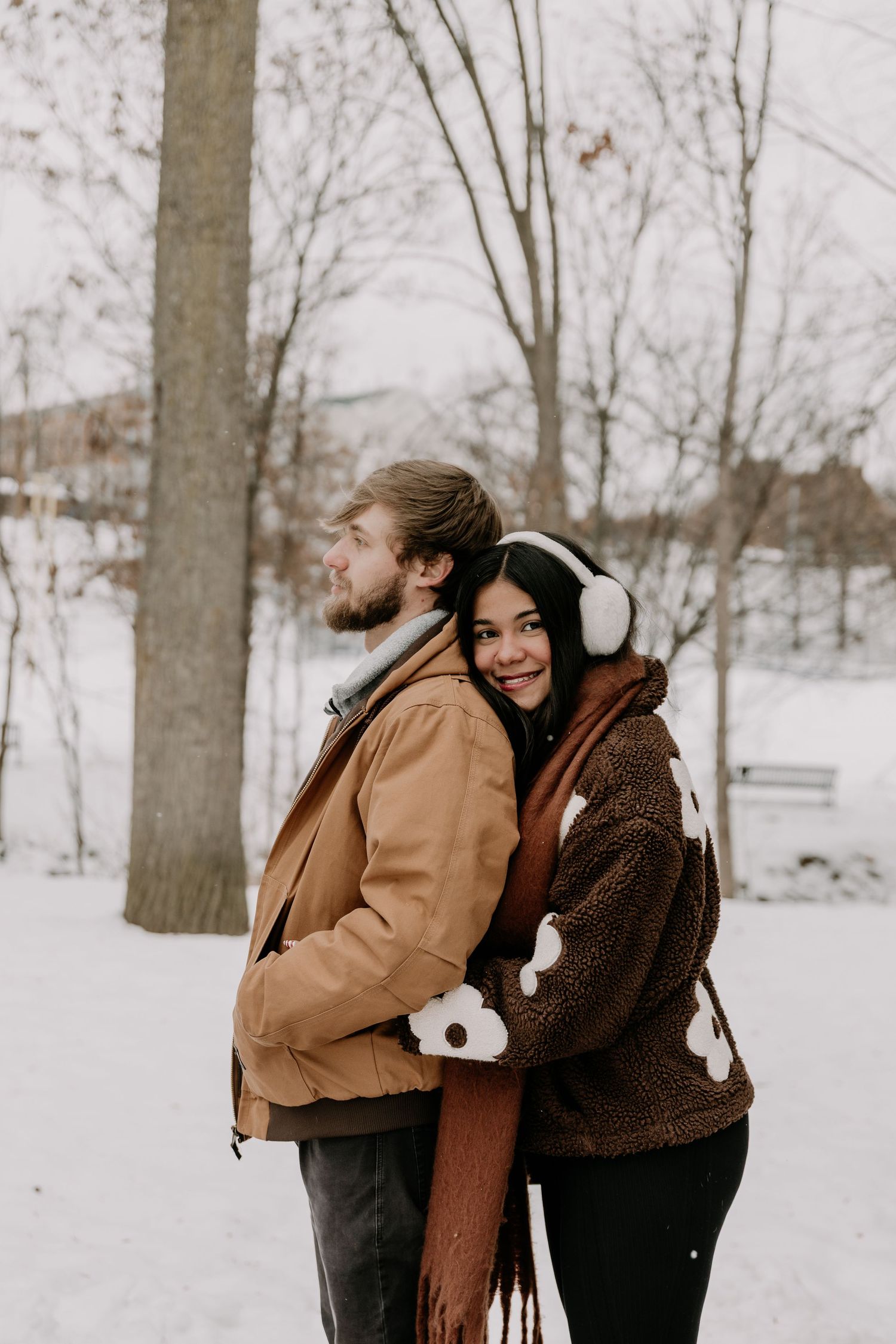 Two people in warm winter coats embrace in a snowy park setting with bare trees in the background.