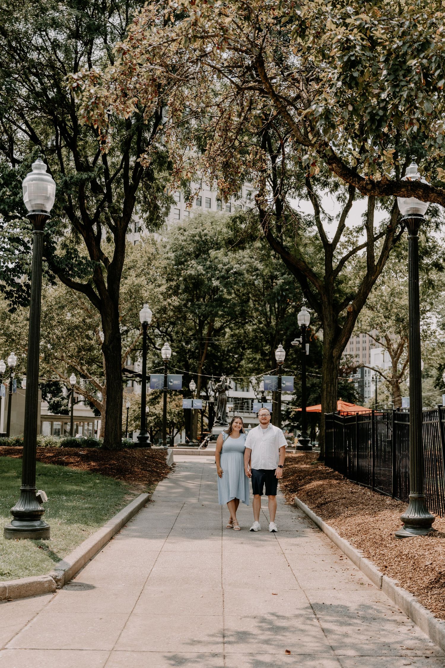 Two people walking together on a tree-lined path with vintage street lamps in an urban park setting on a sunny day.