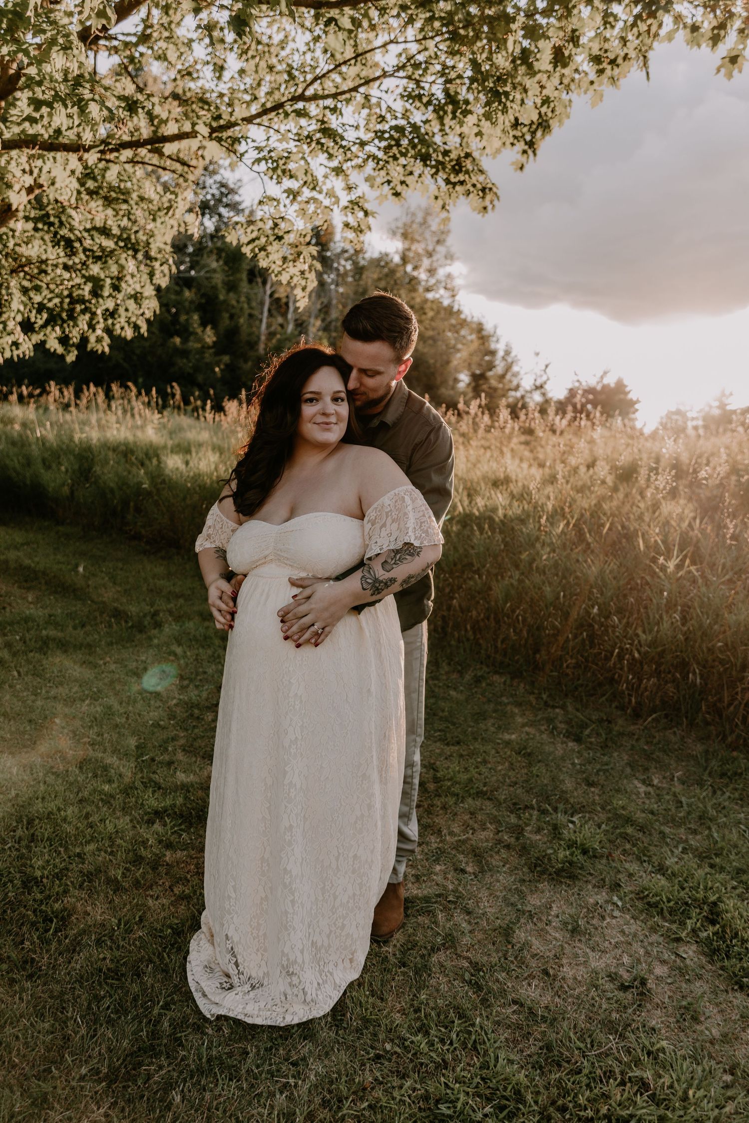 A romantic maternity photo shoot in a sunlit field shows a couple embracing in golden hour lighting.