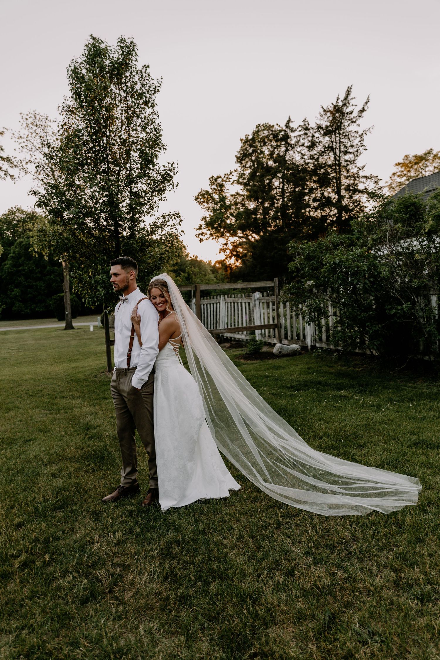 Bride in flowing veil embraces groom in suspenders during golden hour wedding portraits on lush green lawn.