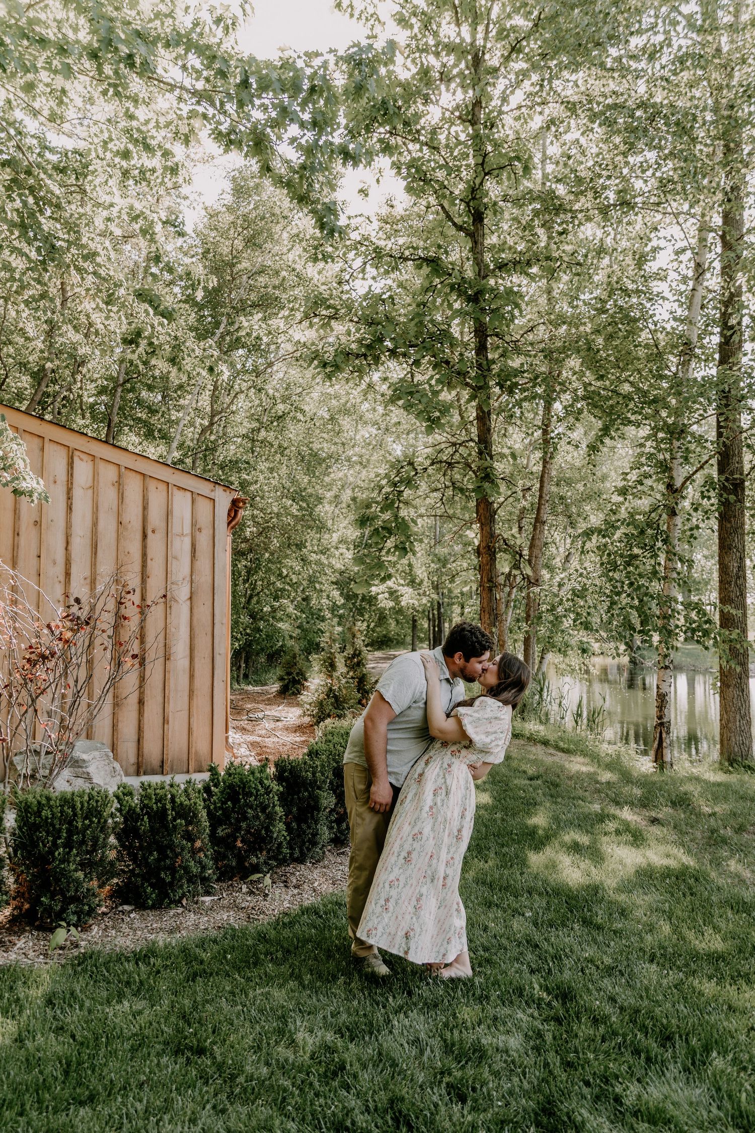 Couple sharing a romantic dip kiss on lush green lawn surrounded by tall trees near a wooden shed.