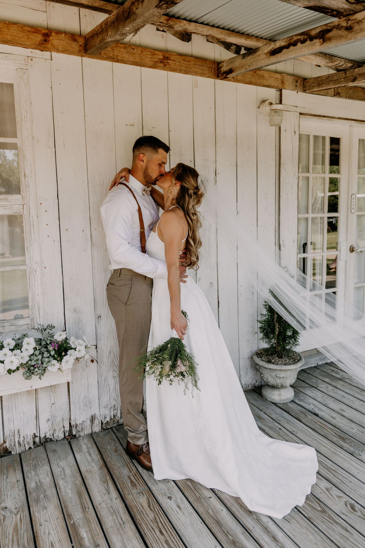 Bride and groom sharing a kiss on a rustic white wooden porch, bride holding a green and white bouquet.