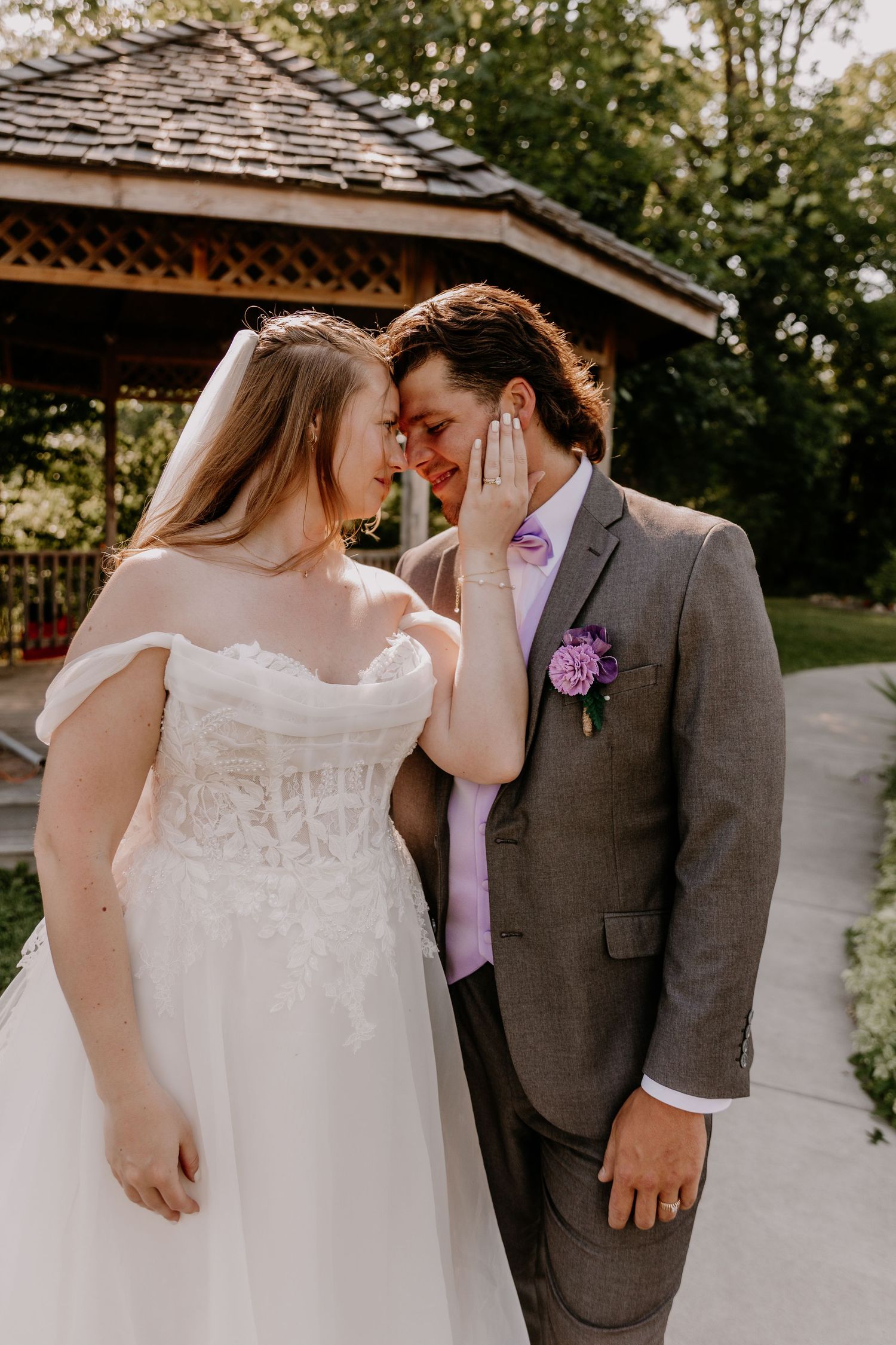 A bride and groom share an intimate forehead touch outdoors, she in a white lace gown, he in a gray suit with purple boutonniere.