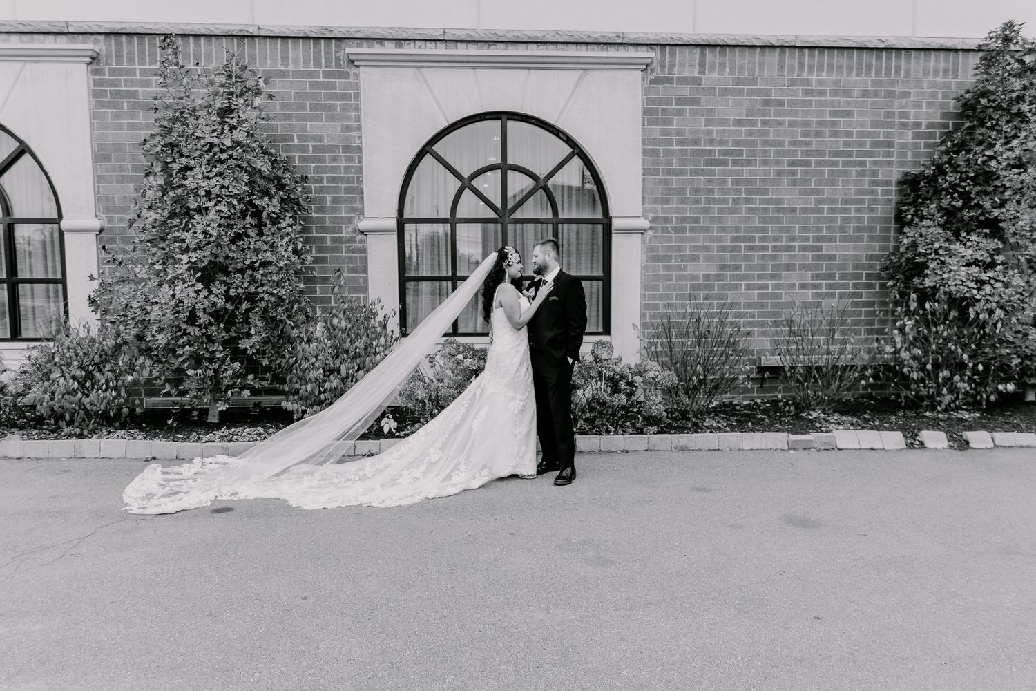Classic black and white portrait of newlyweds against brick wall with arched windows.