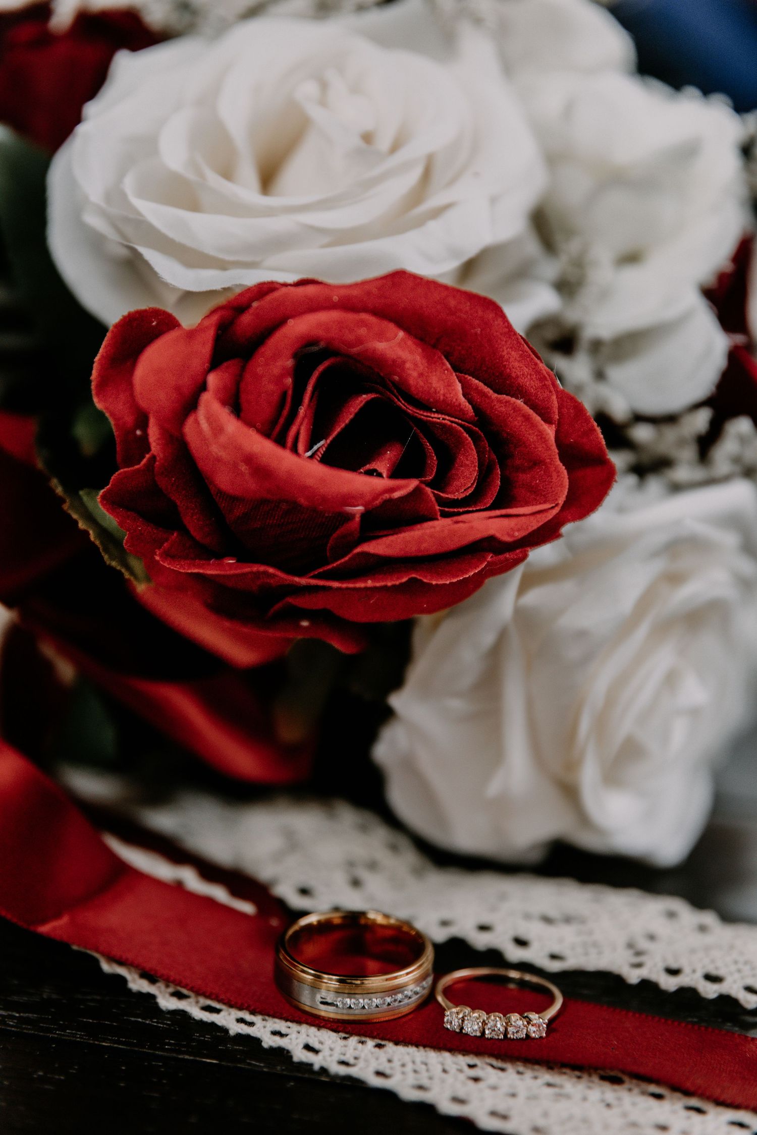 Wedding rings displayed with red and white roses on white lace fabric.